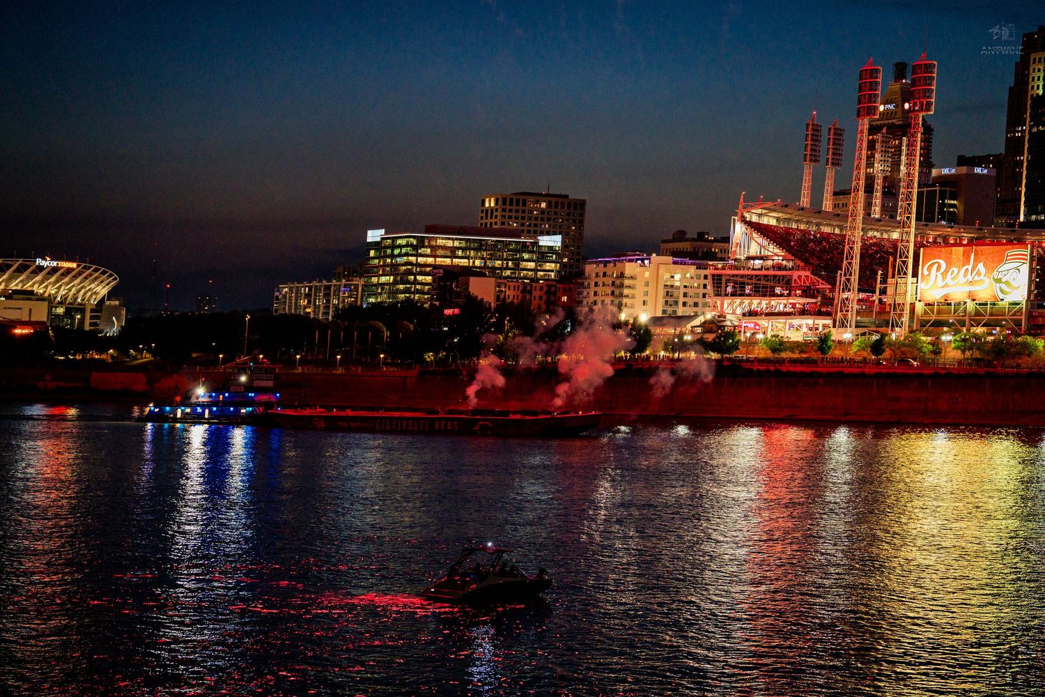 Nighttime cityscape with illuminated stadium and buildings reflecting on a river with a boat in the foreground.