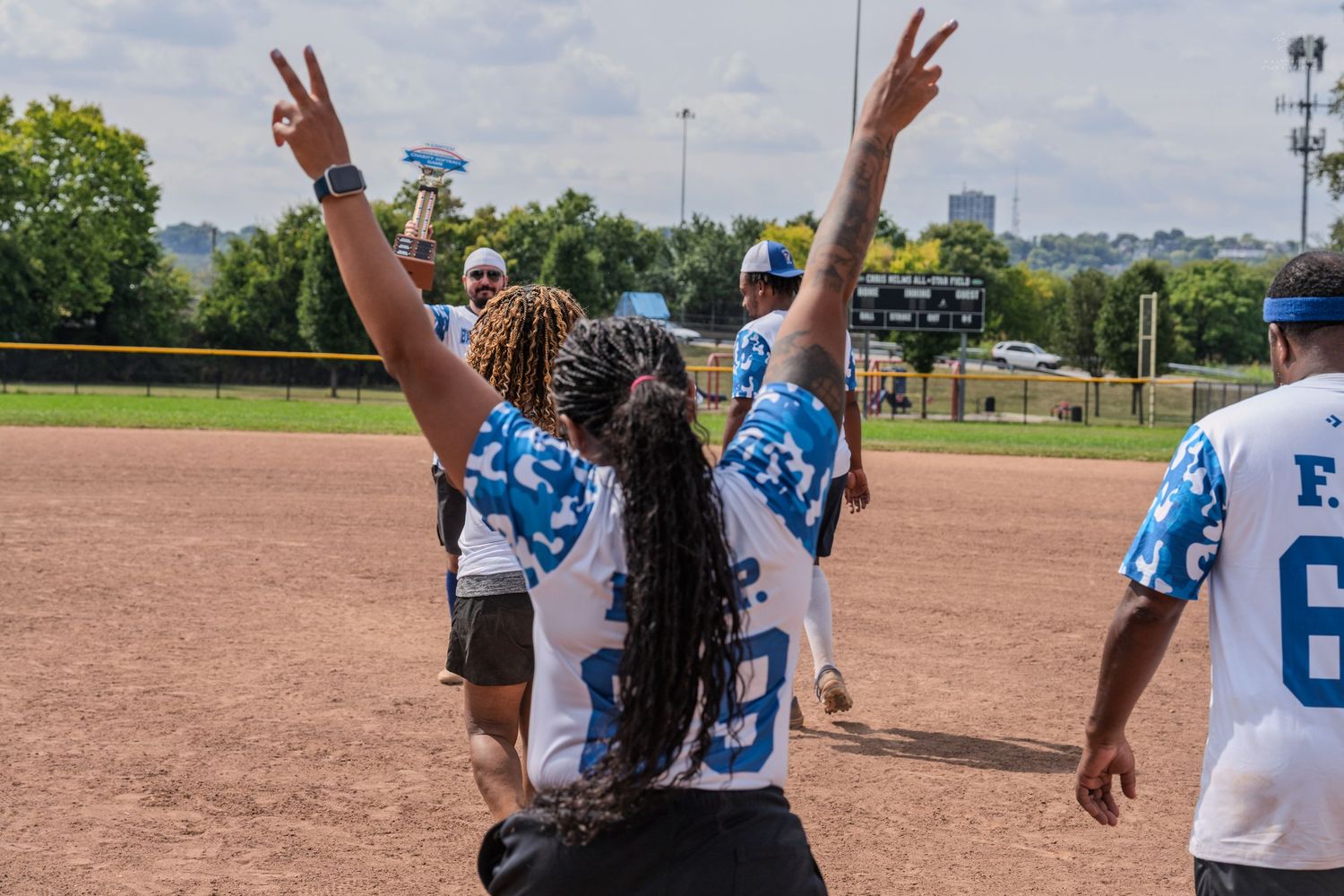Two softball players in blue jerseys raise their arms in victory celebration on a sunny baseball field.