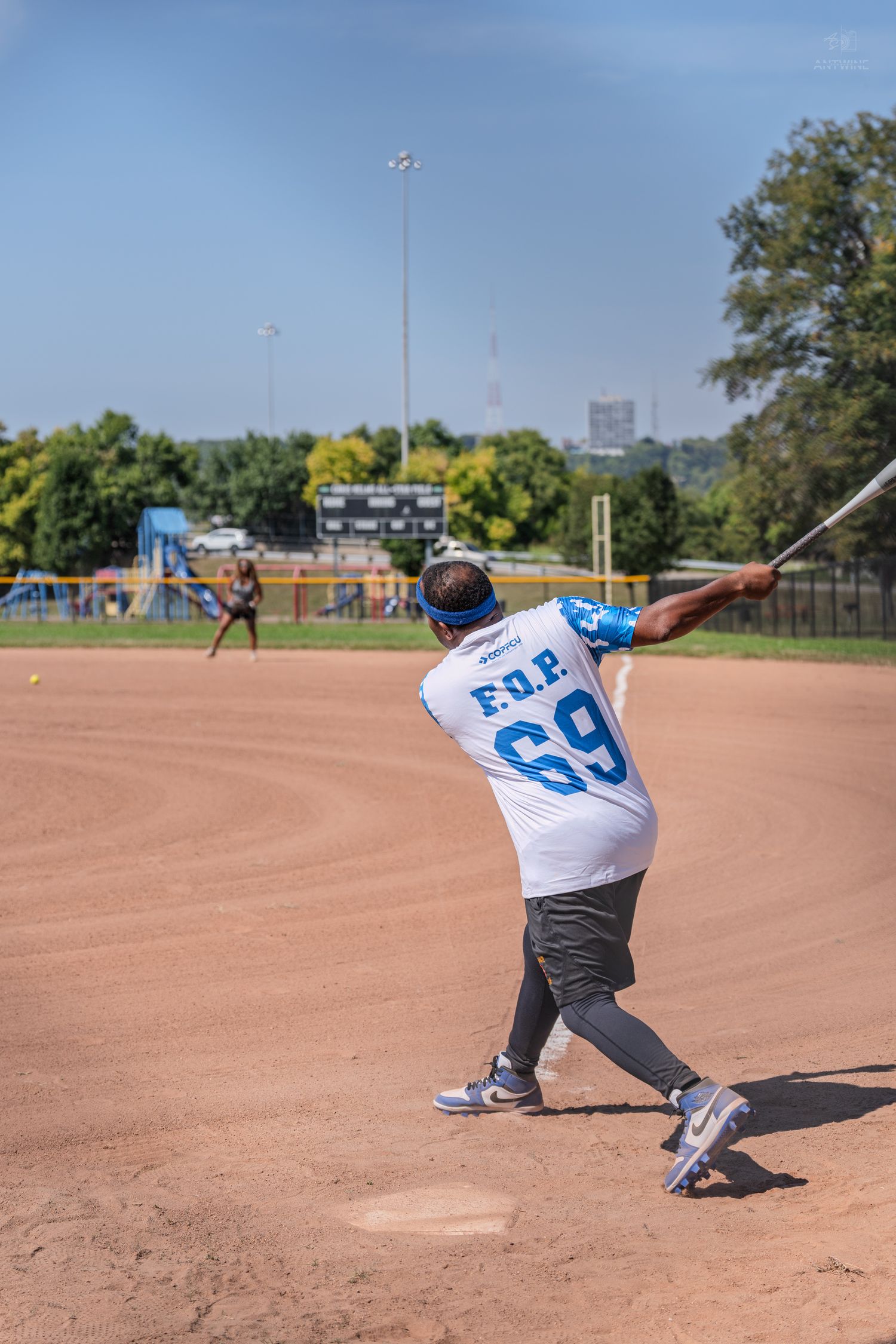 Softball batter in F.O.P. number 69 jersey completing a swing with pitcher visible in background.