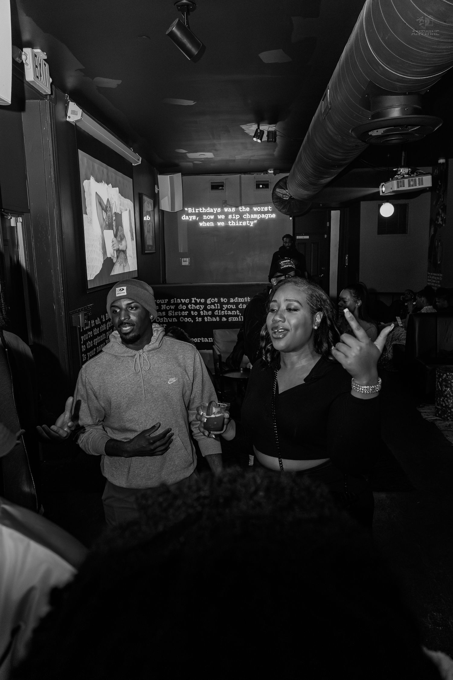 Black and white photo of two people singing karaoke in a dimly lit bar, woman gesturing with microphone.