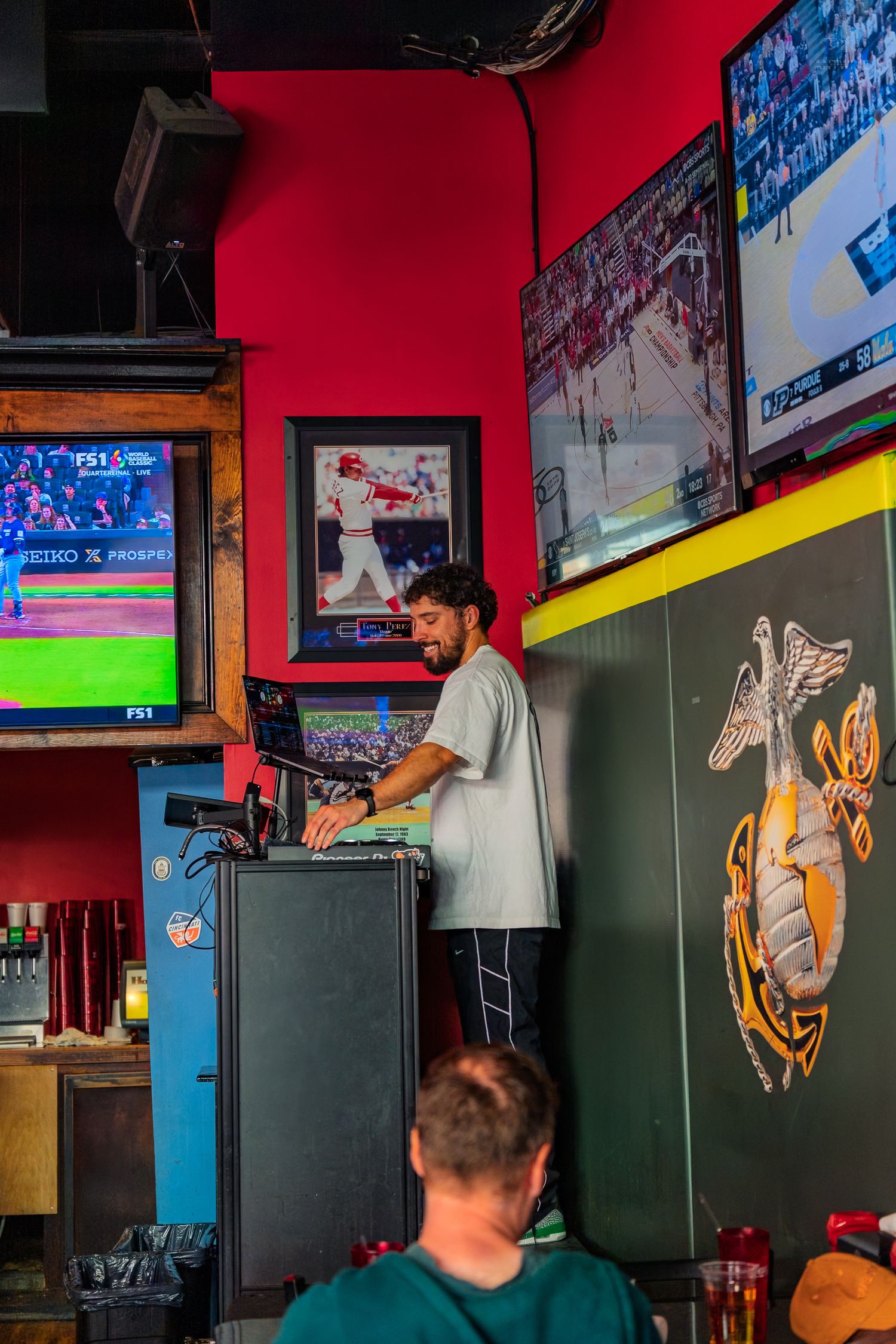 A DJ operates equipment in a lively sports bar with red walls and multiple TVs showing baseball and hockey games.