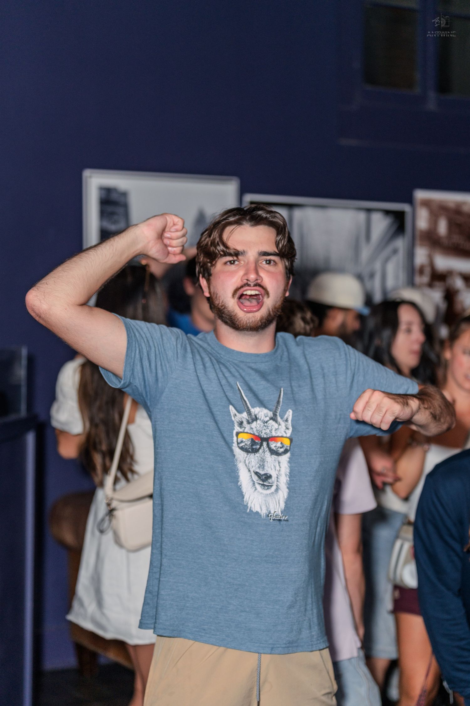 Excited young man in llama graphic tee flexes arms at a lively indoor social gathering.
