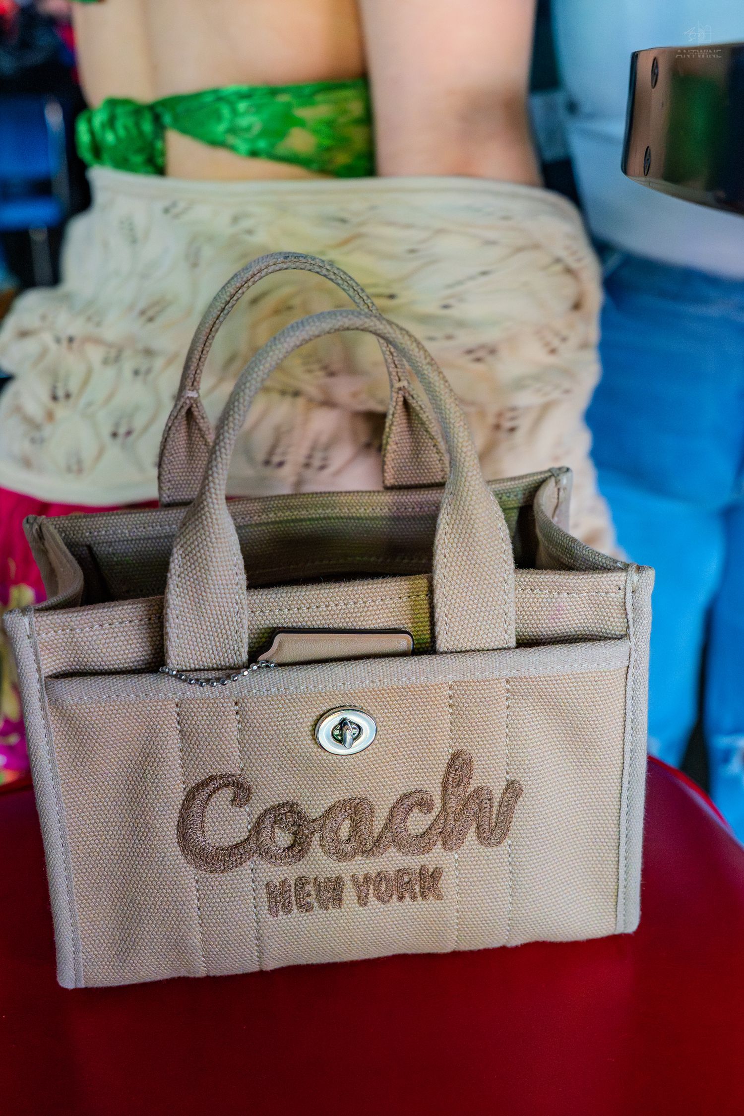 Beige Coach New York canvas tote bag with embroidered logo and top handles displayed on a red surface.