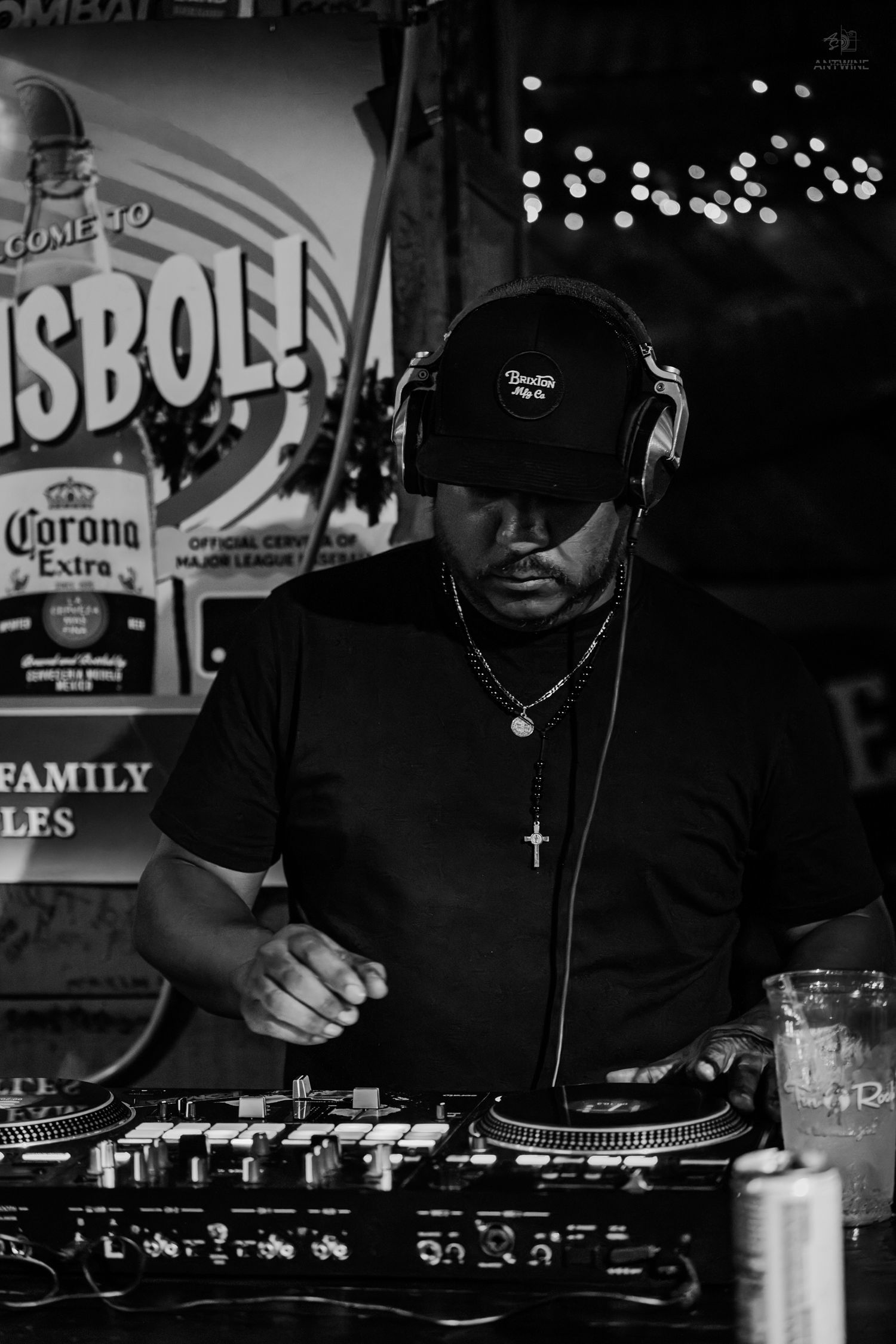 Black and white photo of a DJ wearing headphones and a cross necklace, mixing at turntables near a Corona Extra sign.