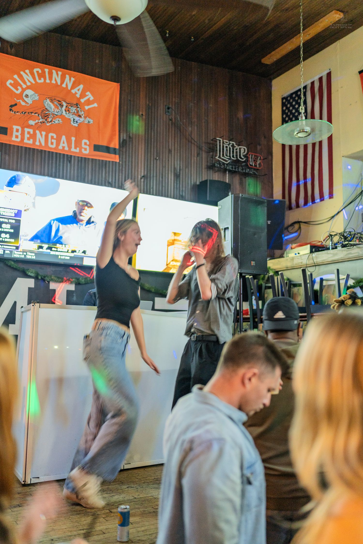 Two women dancing near a DJ booth inside a bar decorated with a Cincinnati Bengals flag.