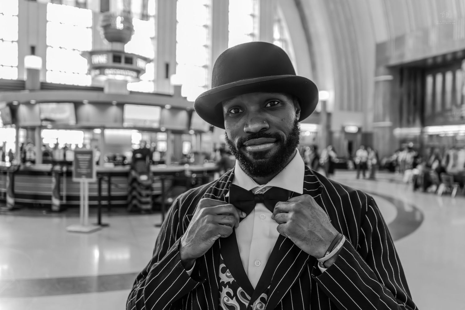 Stylish Black man in pinstripe suit and bowler hat adjusting bow tie in a busy train station, black and white photo.