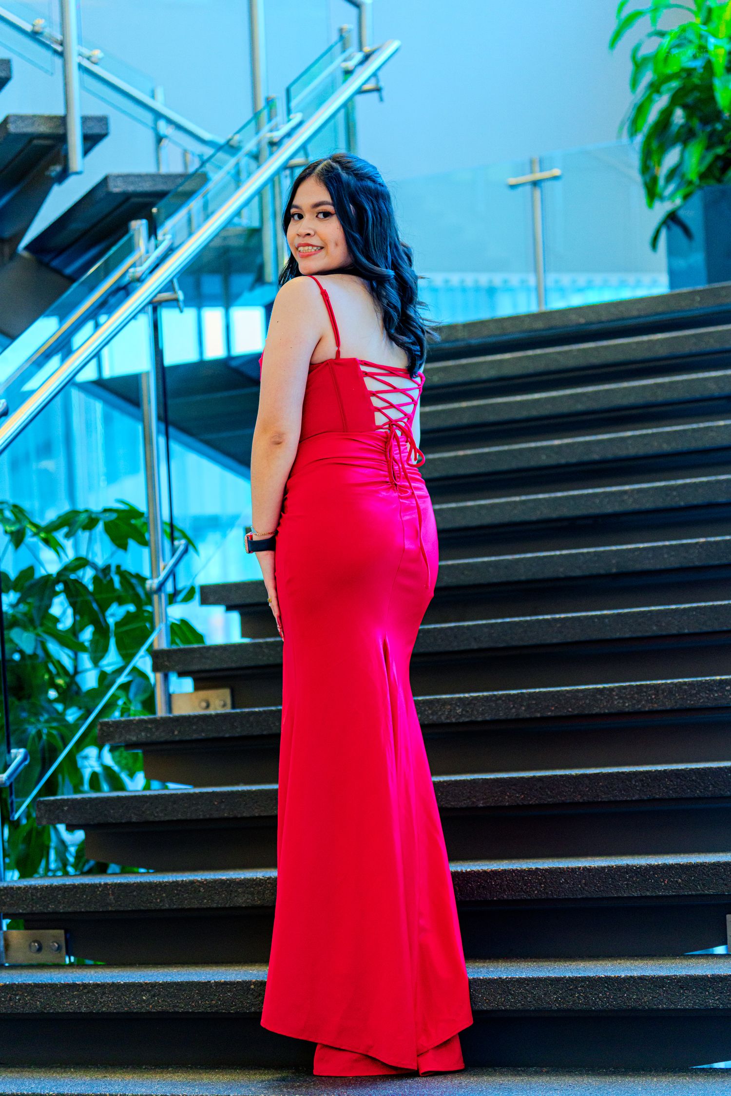 Woman in red lace-up back formal gown posing on modern staircase from behind.
