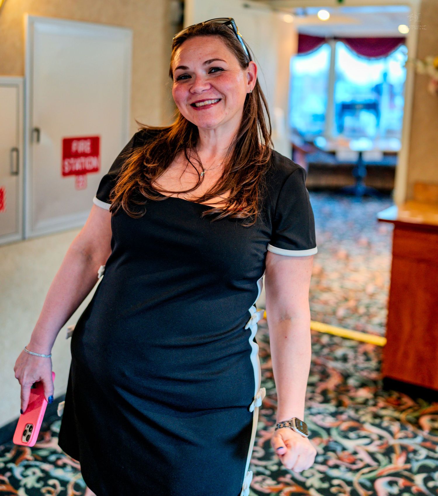 Smiling woman in black dress walking through an elegant carpeted hotel hallway holding a pink phone.