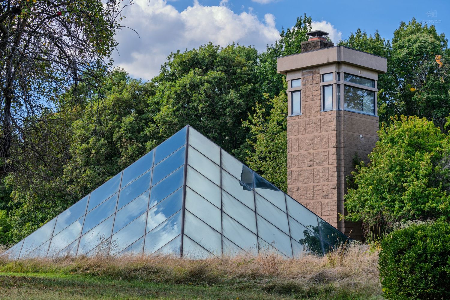 Glass pyramid structure beside a brick tower surrounded by lush green trees under a partly cloudy sky.