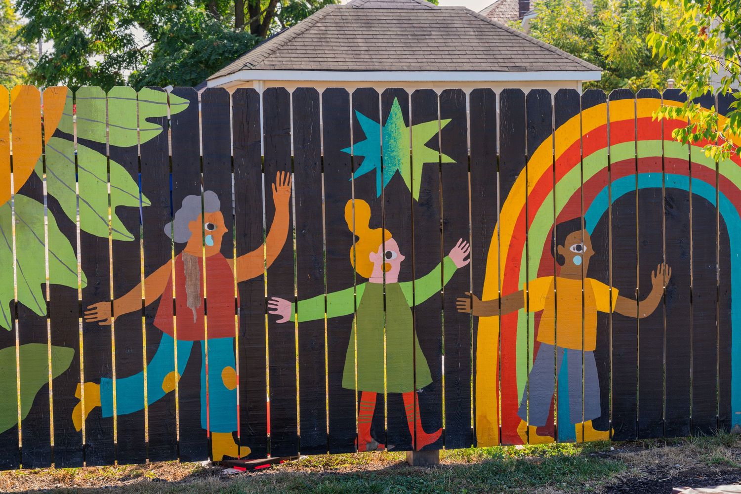 Colorful mural on a wooden fence featuring three figures dancing near a rainbow and star on a dark background.