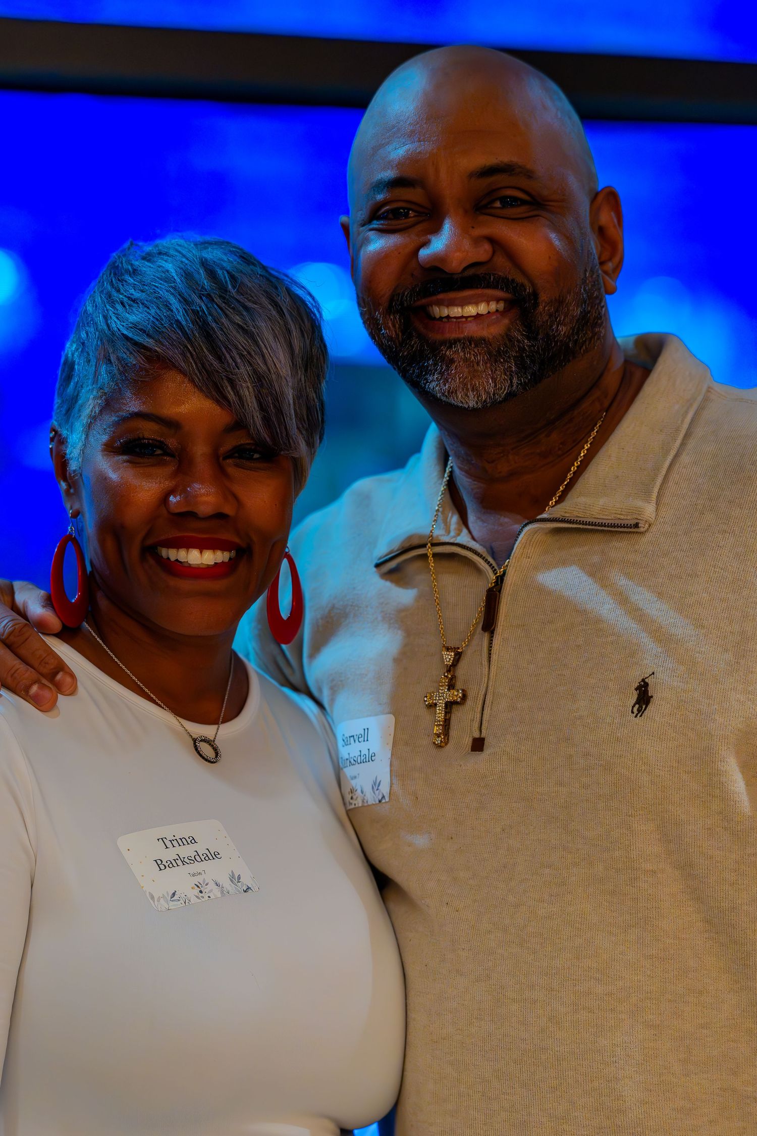 Two smiling people wearing name tags pose together at an event, with a blue-lit background behind them.