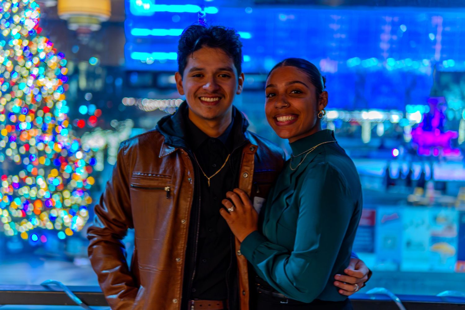 Smiling couple posing together near a decorated Christmas tree with colorful bokeh lights at night.