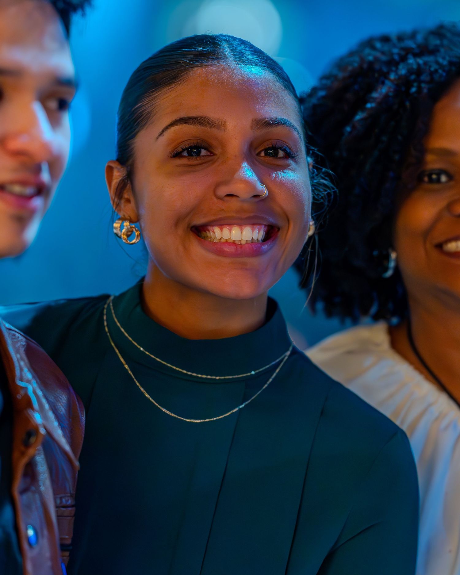 Smiling young woman with gold earrings and layered necklaces stands with friends in blue-lit venue.