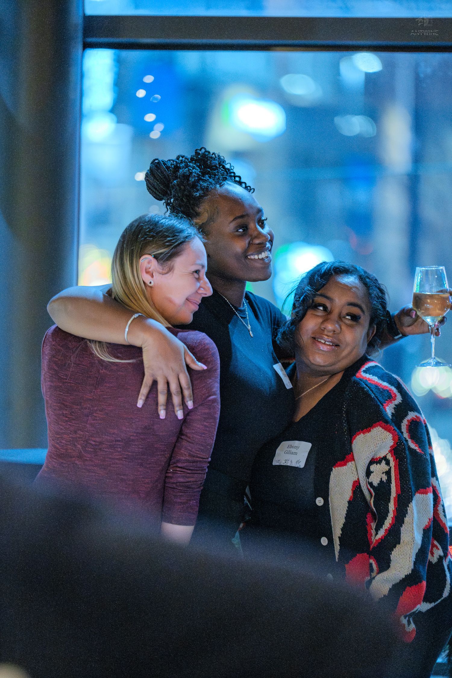 Three women smiling and posing together at a social event, one holding a glass of wine.