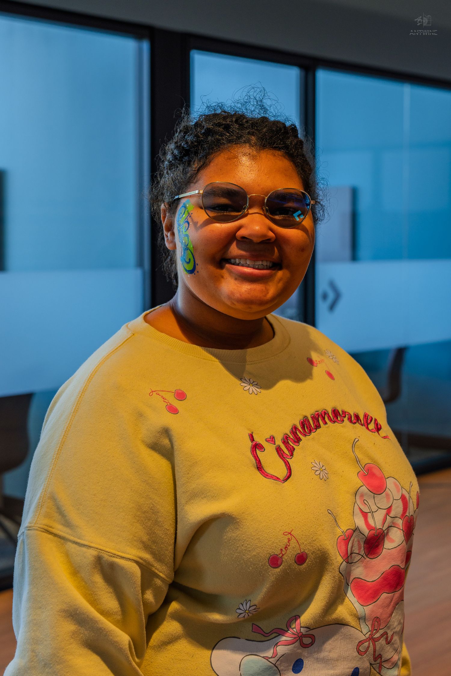 A smiling young woman wearing sunglasses and a yellow sweatshirt shows off colorful face paint designs.
