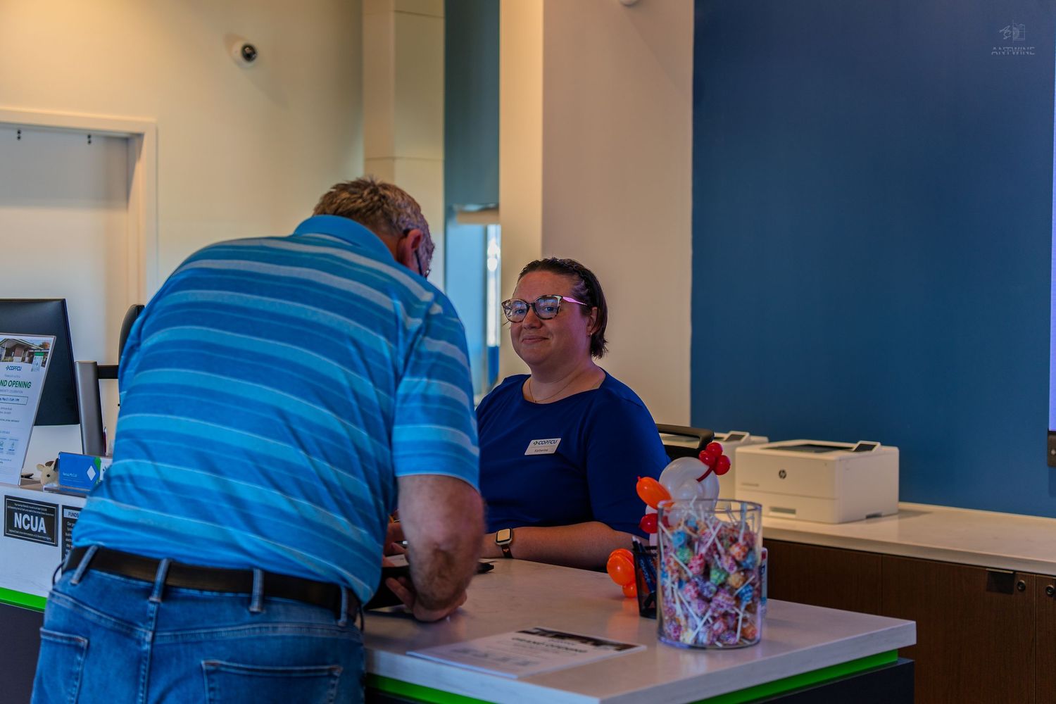 Smiling credit union employee assists a customer at a welcome registration desk during a grand opening.