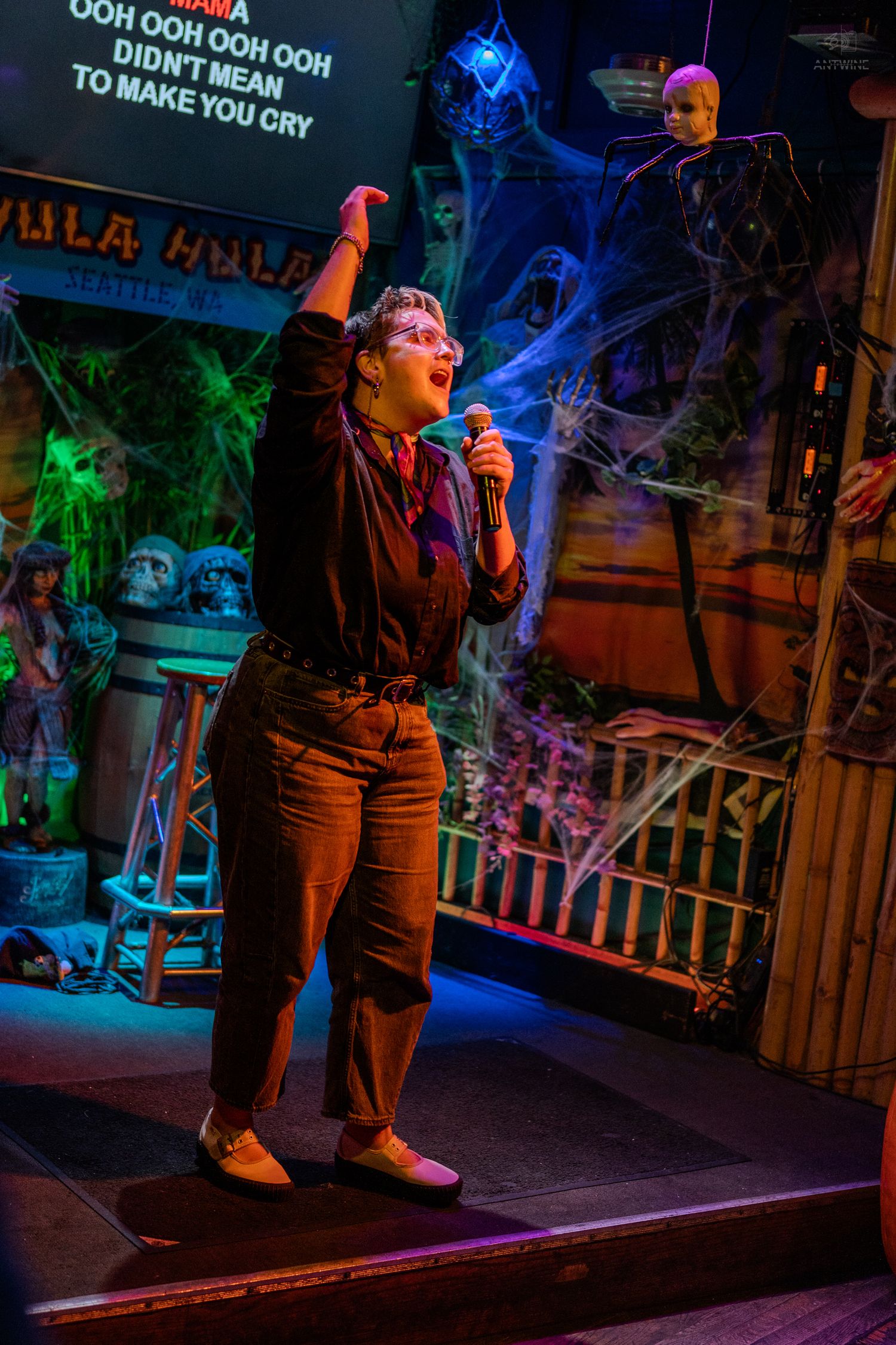 Woman singing karaoke on Halloween-decorated stage at Hula Hula bar in Seattle, WA, arm raised holding microphone.