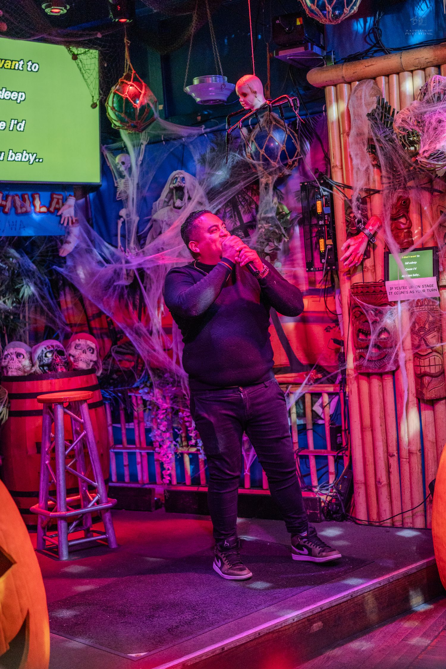 Man passionately singing karaoke at Halloween-decorated bar with cobwebs, skulls, and colorful lighting.