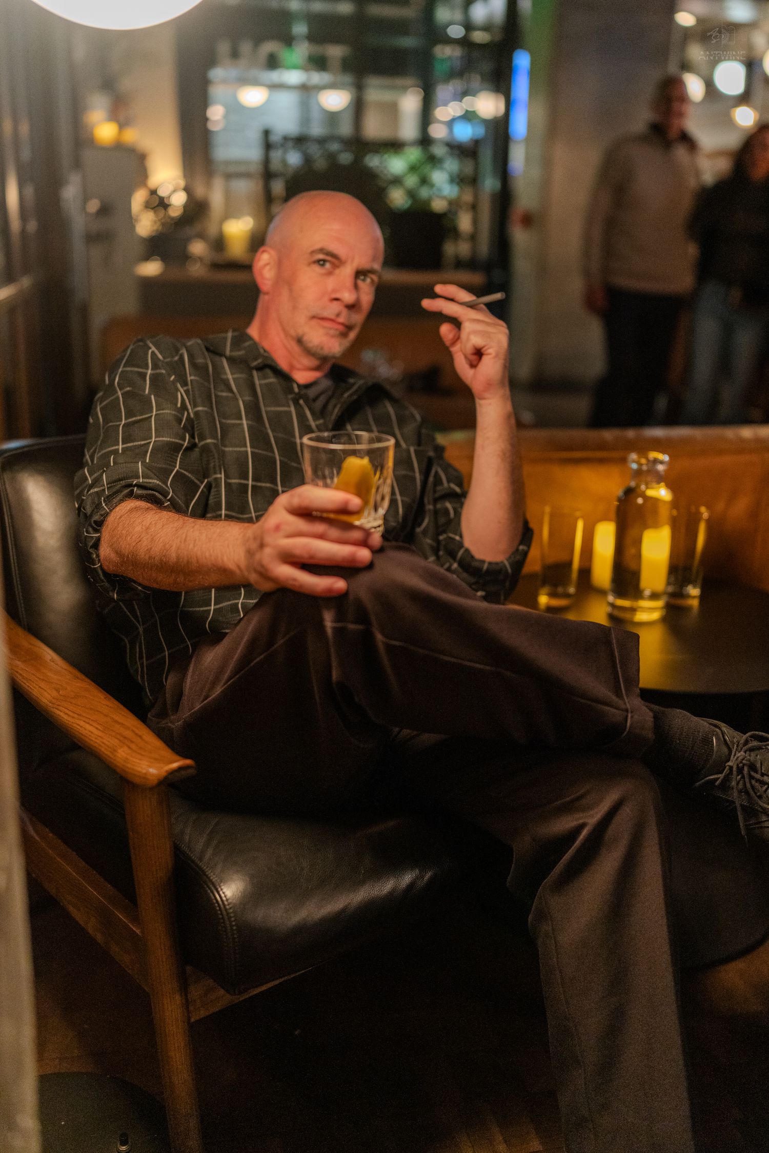 A bald man relaxes in a leather chair holding a beer and cigarette in a dimly lit bar.