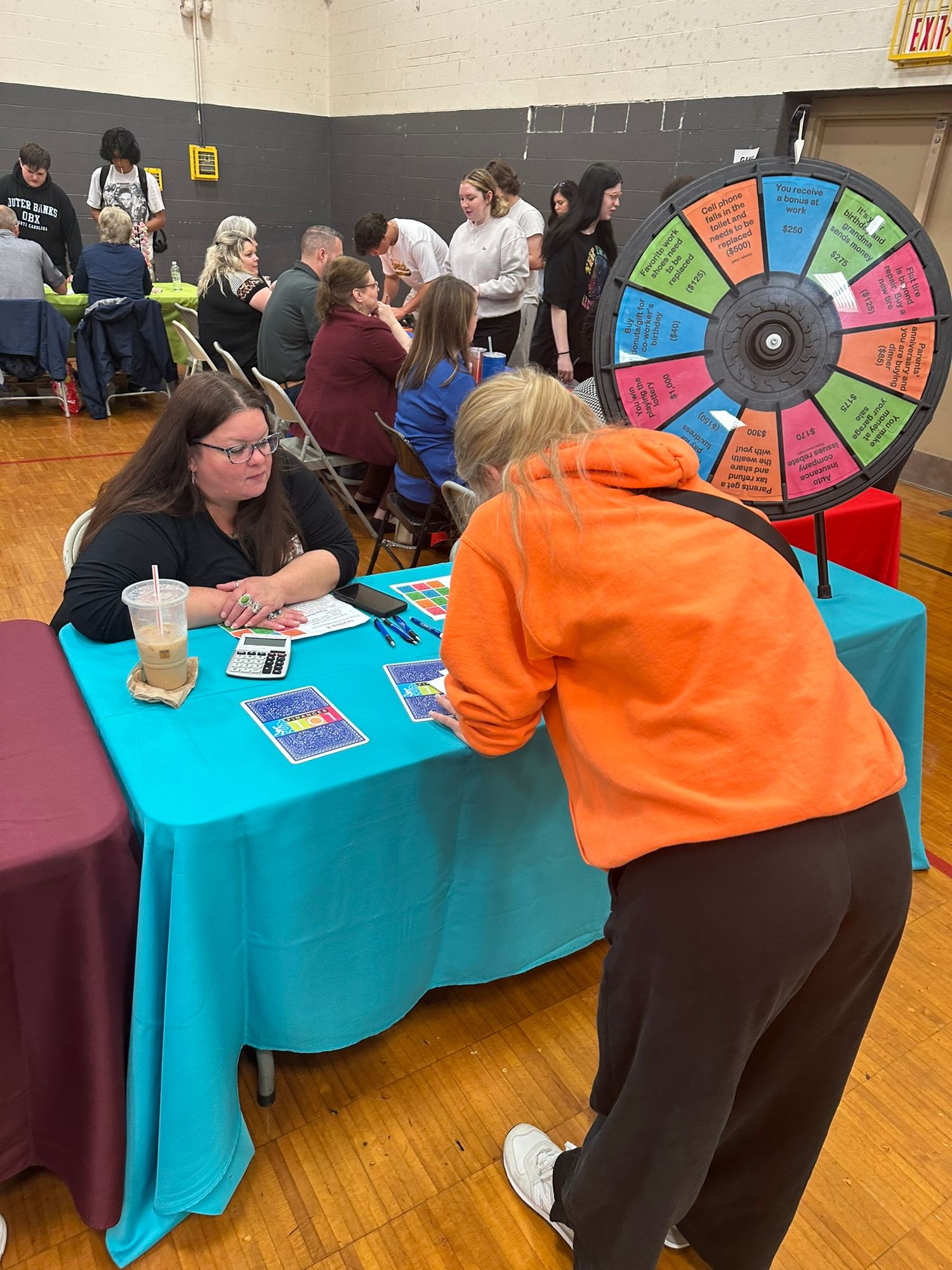 Students interact at a colorful booth with a prize wheel during a school fair event in a gymnasium.
