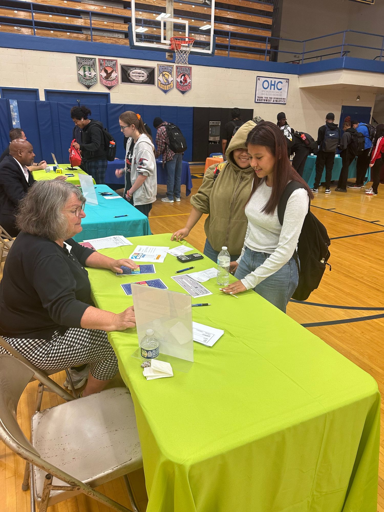 Two students interact with a seated representative at a lime green table during a school gymnasium career fair event.