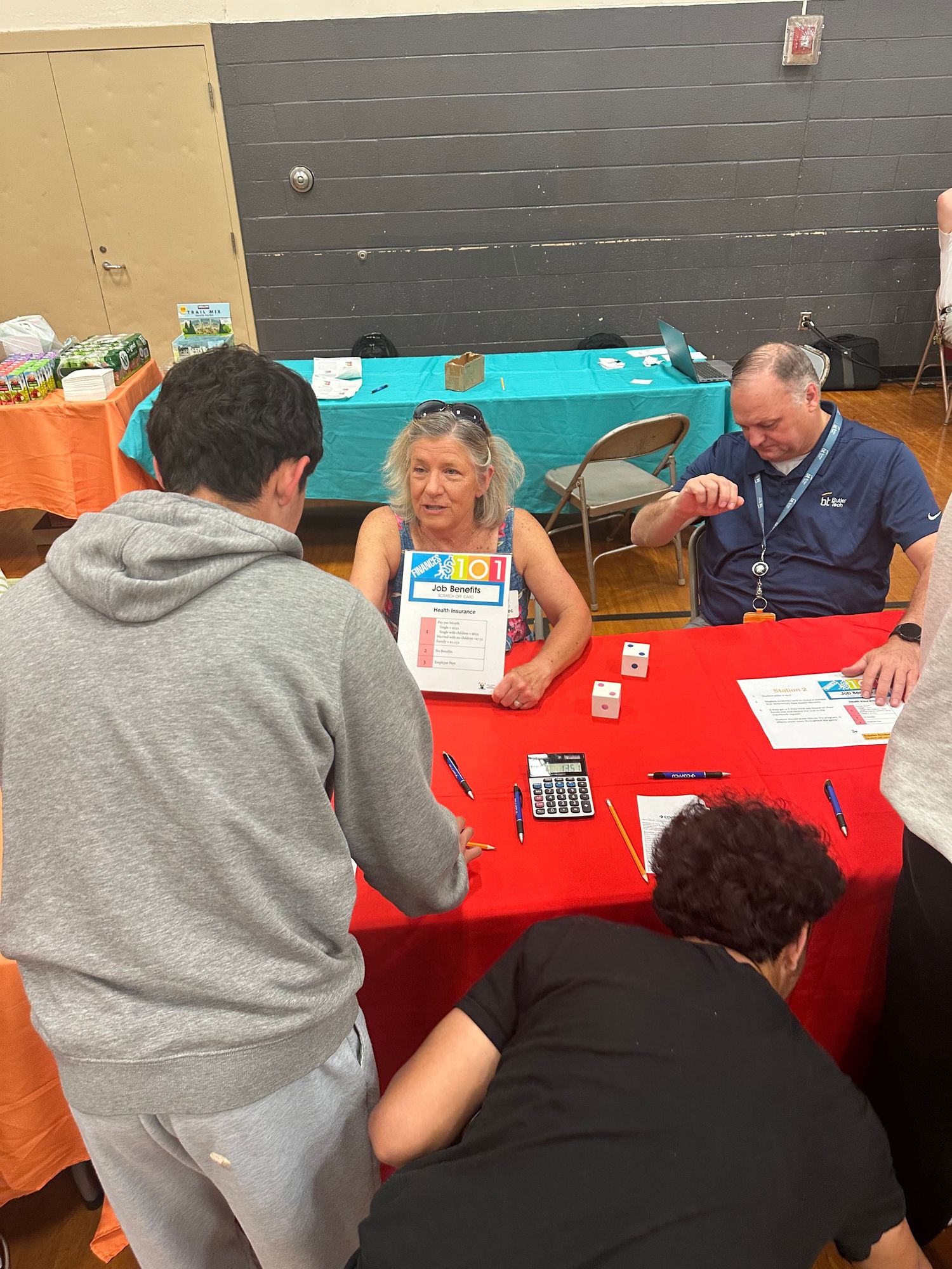 People gathered around a red table at a community event, reviewing documents with organizers in a gymnasium.