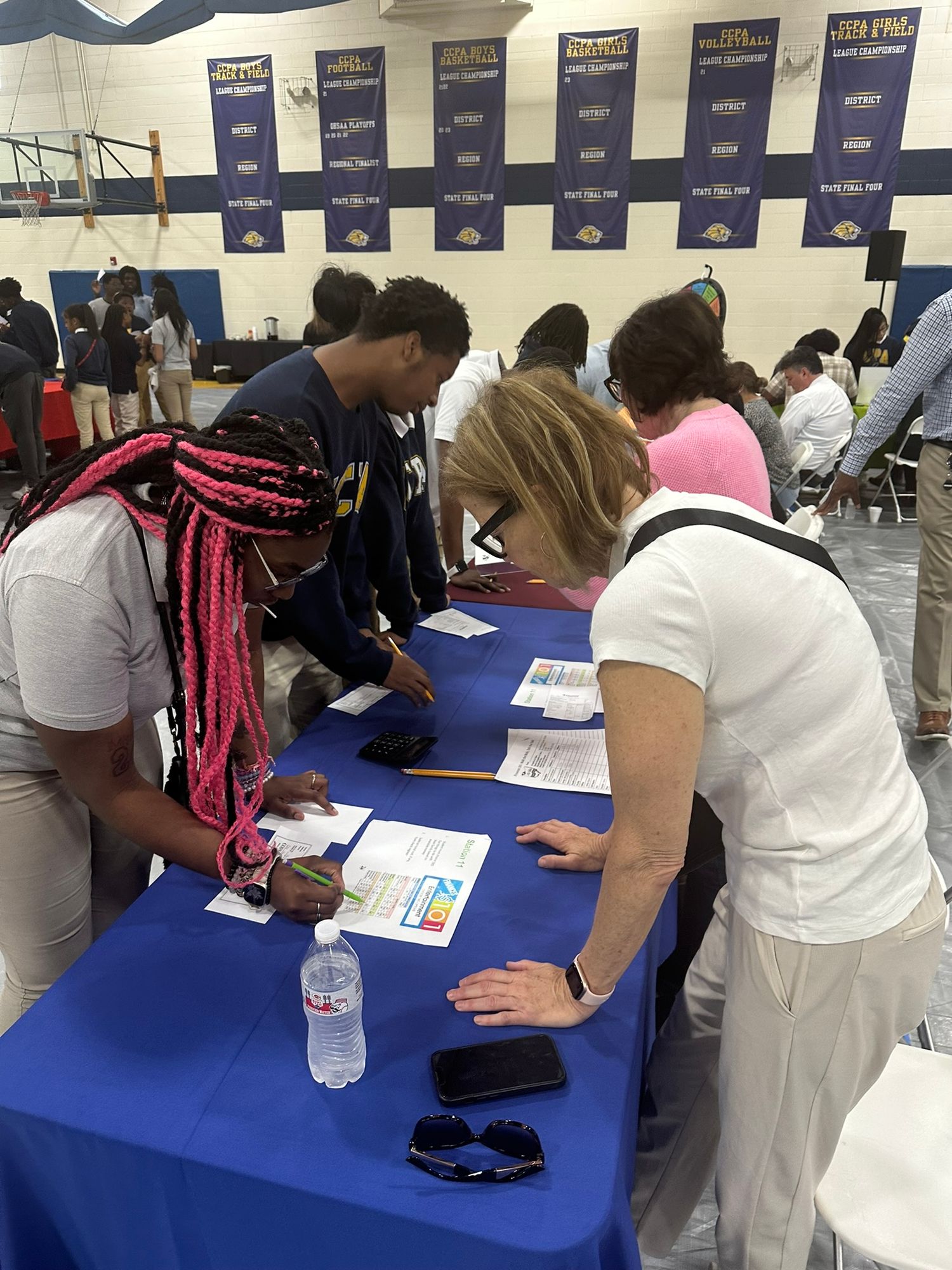 Students and adults signing documents at a blue table during a school gymnasium event or college fair.