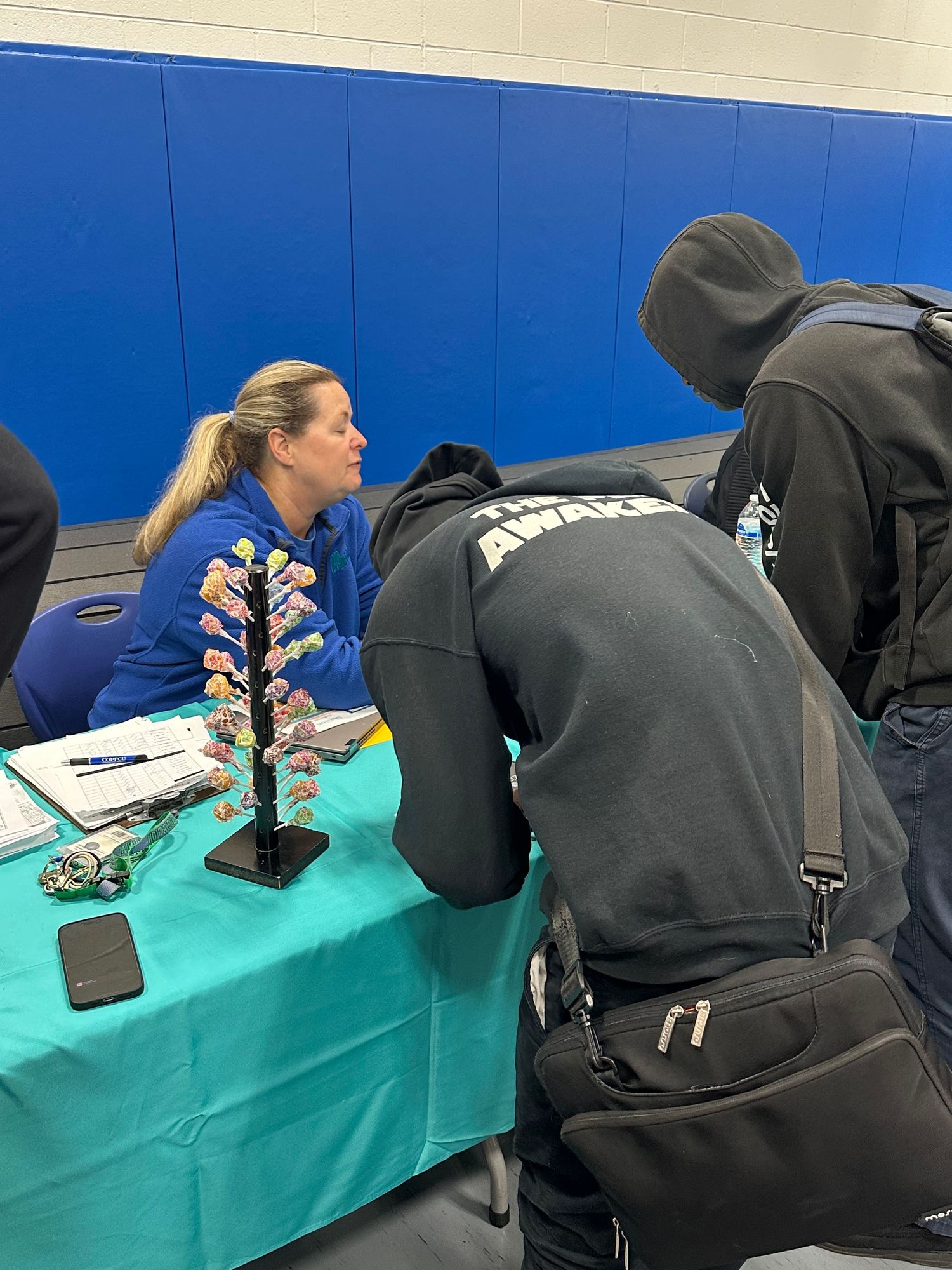 Woman in blue hoodie at teal table speaks with visitor at indoor school gymnasium event or job fair.