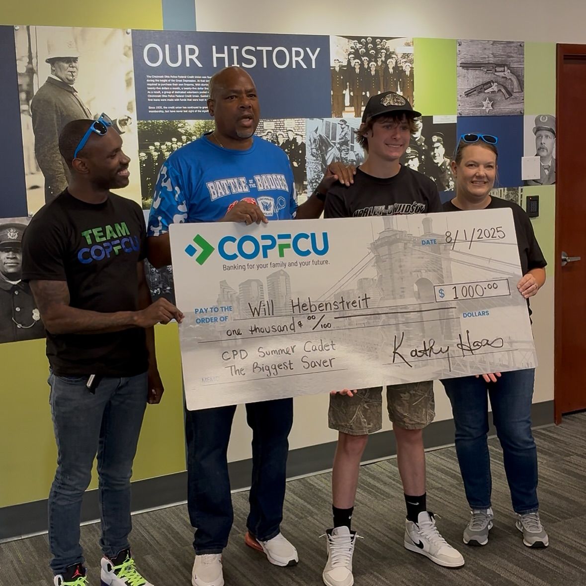 Four people holding a large ceremonial check from COPFCU made out to Will Hebenstreit in front of an Our History wall.