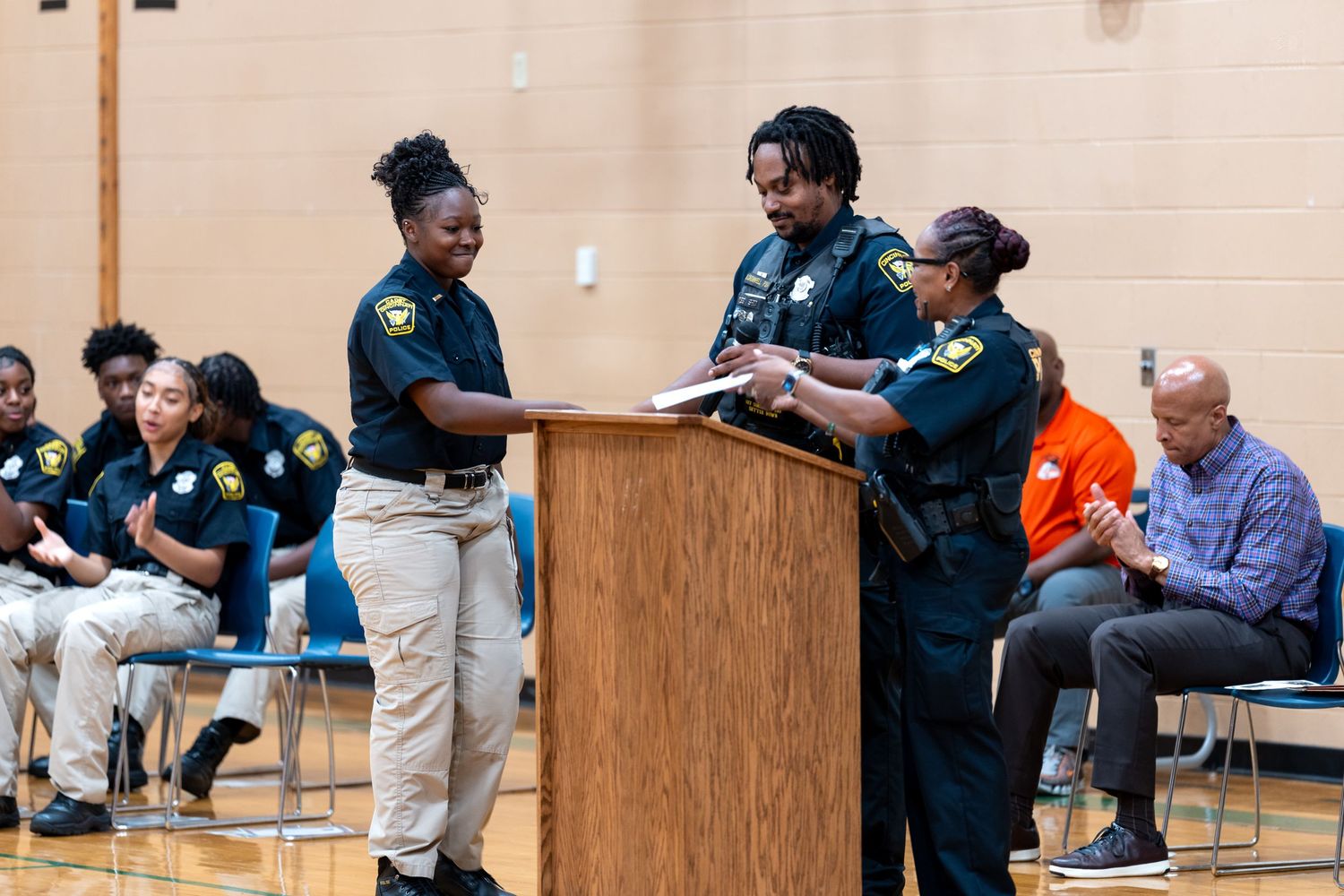 Police officers in uniform stand at a podium during an indoor ceremony in a school gymnasium.