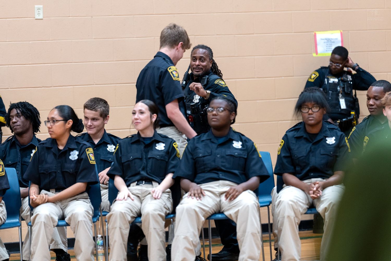 Group of young police cadets in uniform sitting together in a gymnasium during an official ceremony or event.