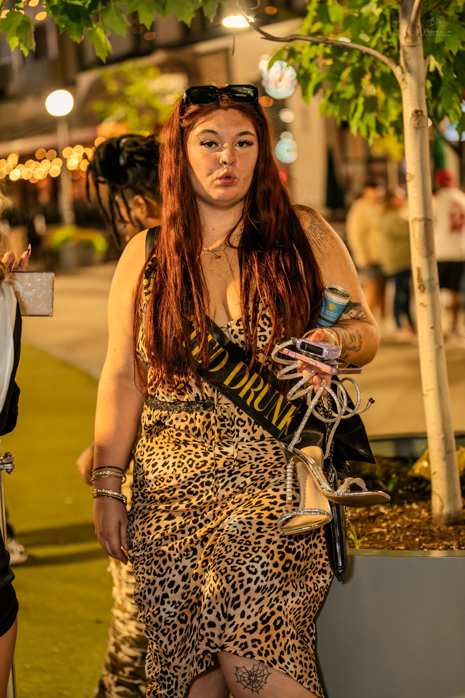 Young woman in leopard print dress with sash poses outdoors at night, holding heels and a drink can.