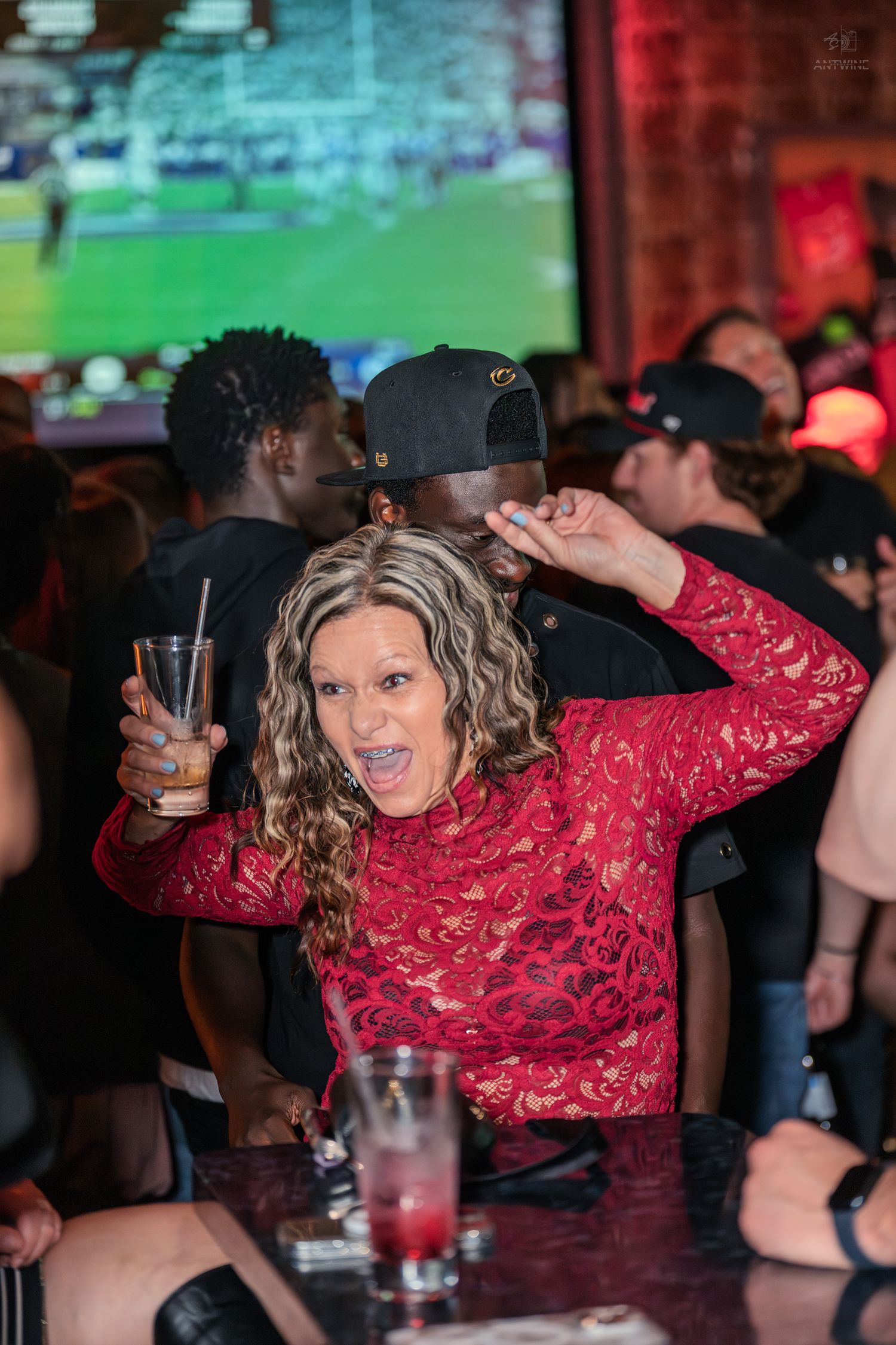 Excited woman in red lace top holding a drink at a lively sports bar nightclub event.