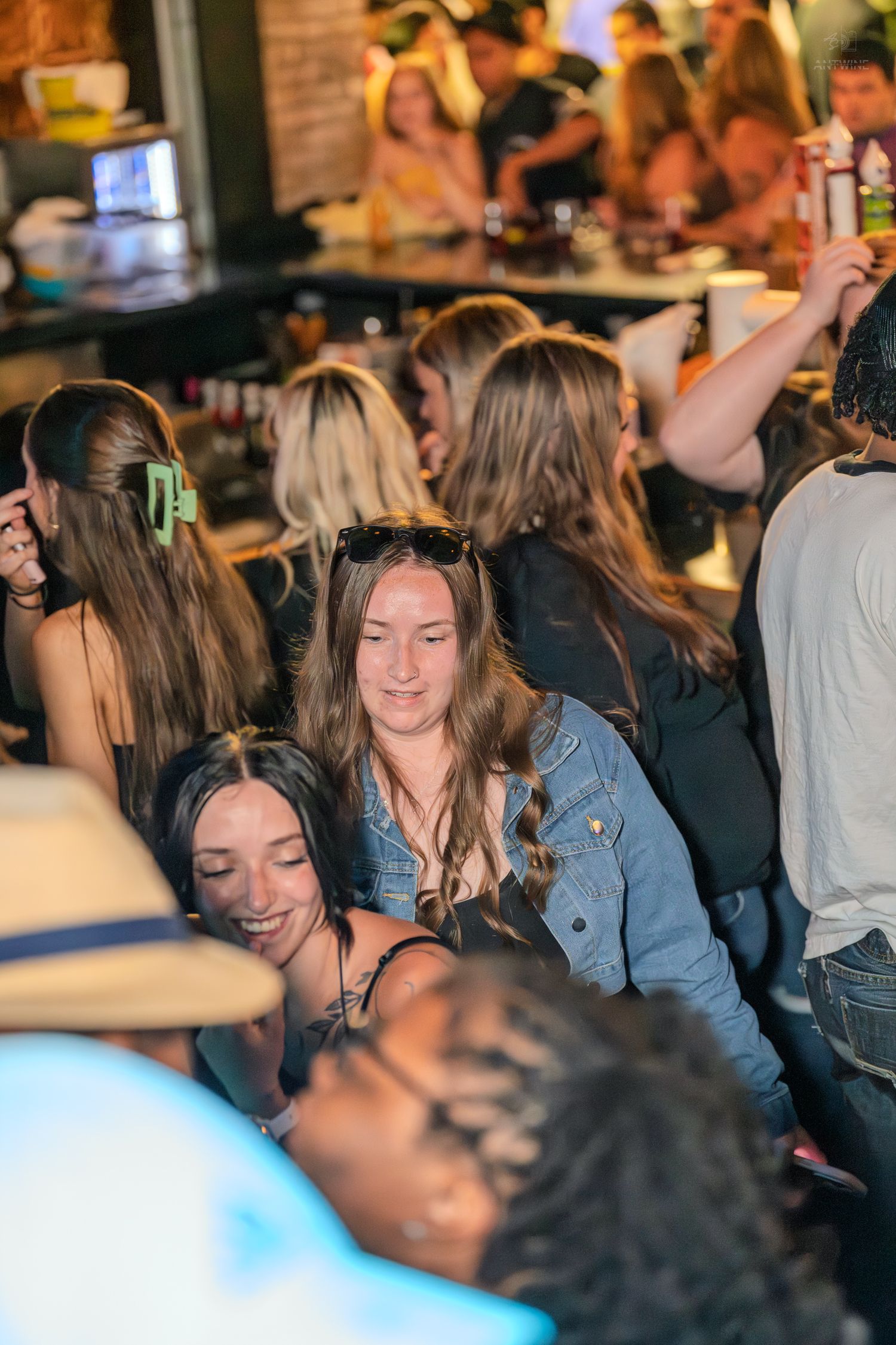 Woman with sunglasses on head smiles in a crowded bar while surrounded by friends in a lively nightlife setting.
