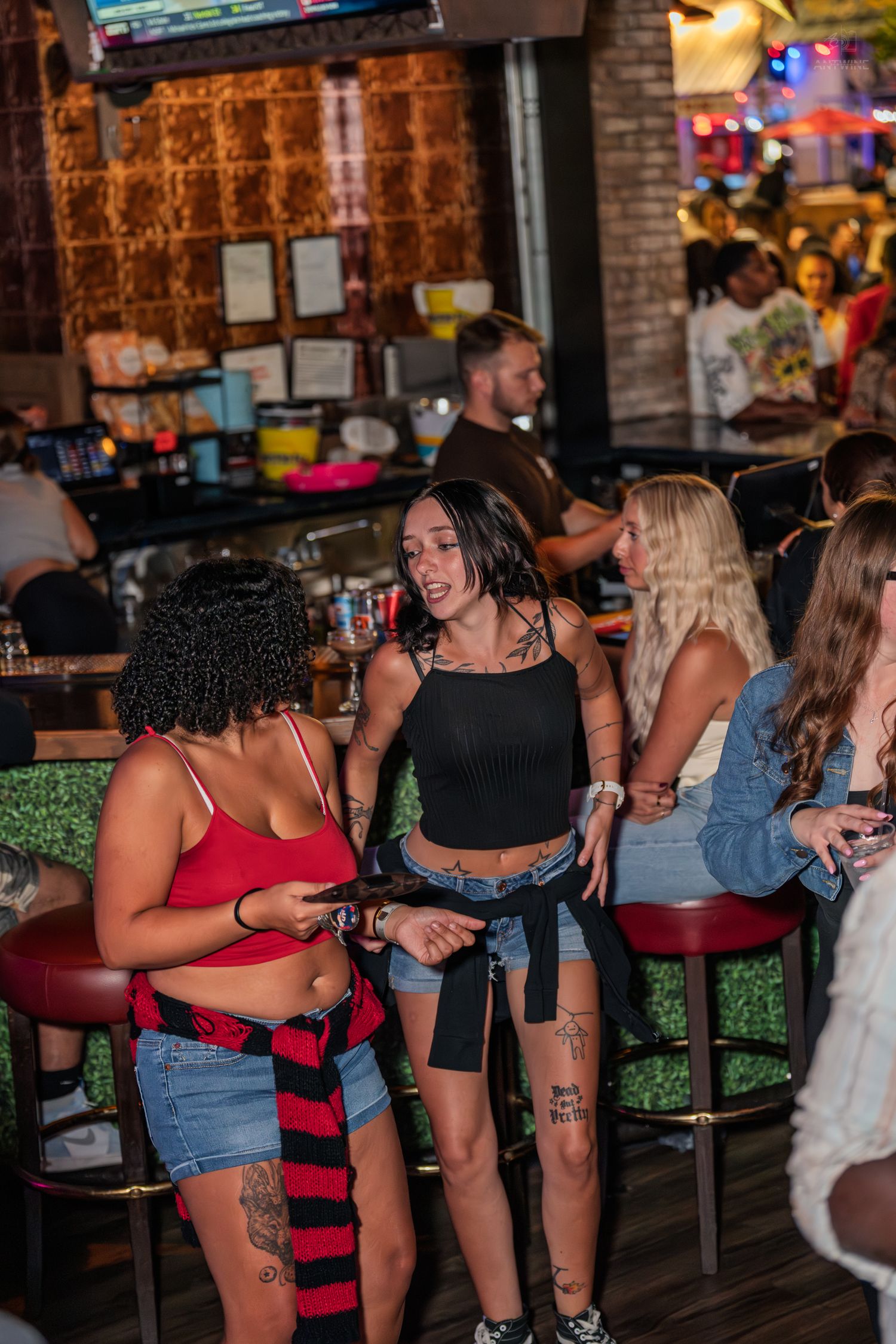 Group of tattooed women socializing and laughing at a lively bar with red barstools.