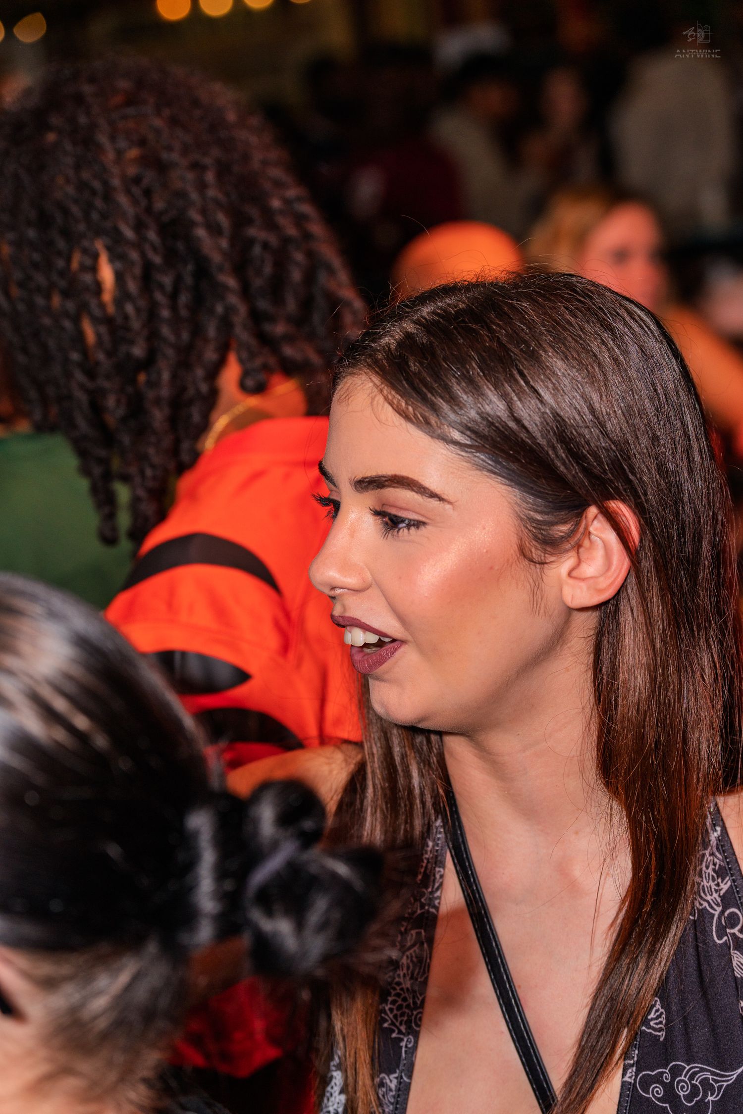 Pretty brunette woman in profile talking to someone at a crowded nightclub event.