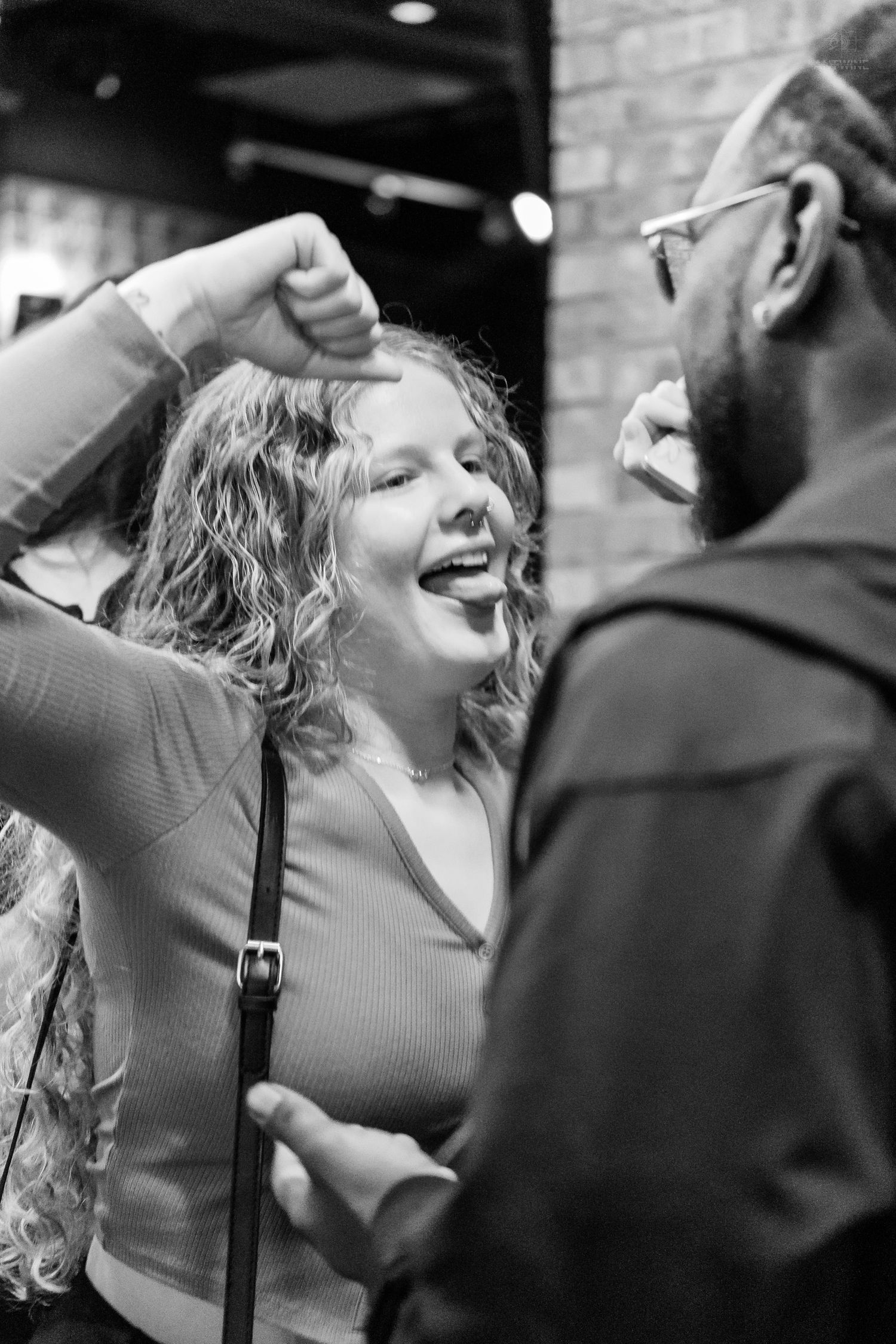 A curly-haired woman laughs and dances while talking to a man outside a brick building.