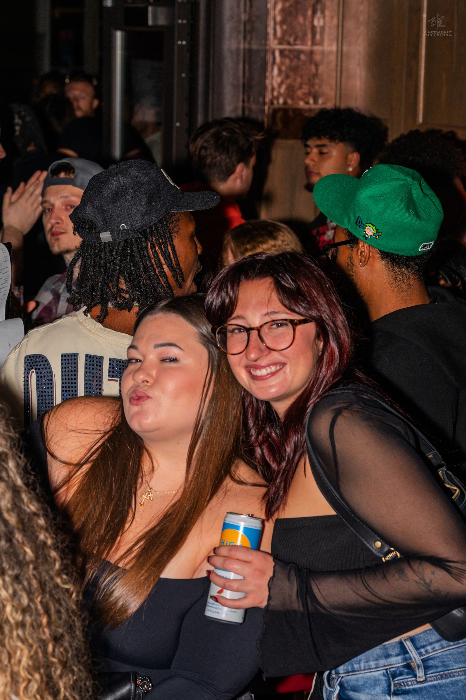 Two women smiling at a crowded nightclub, one holding a canned drink.