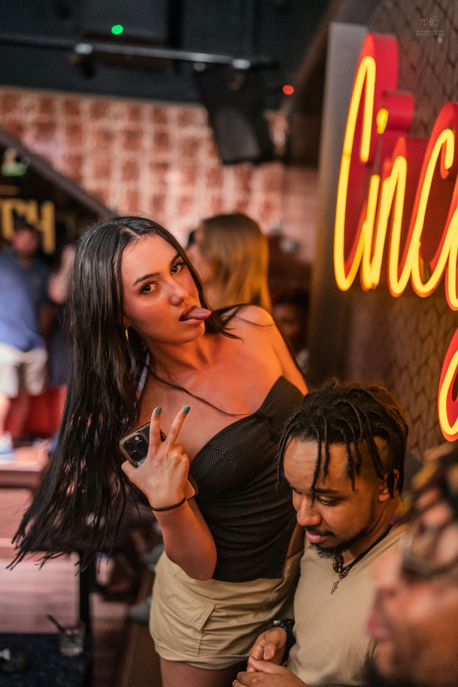 Woman with long dark hair making peace sign gesture at a neon-lit nightclub venue.