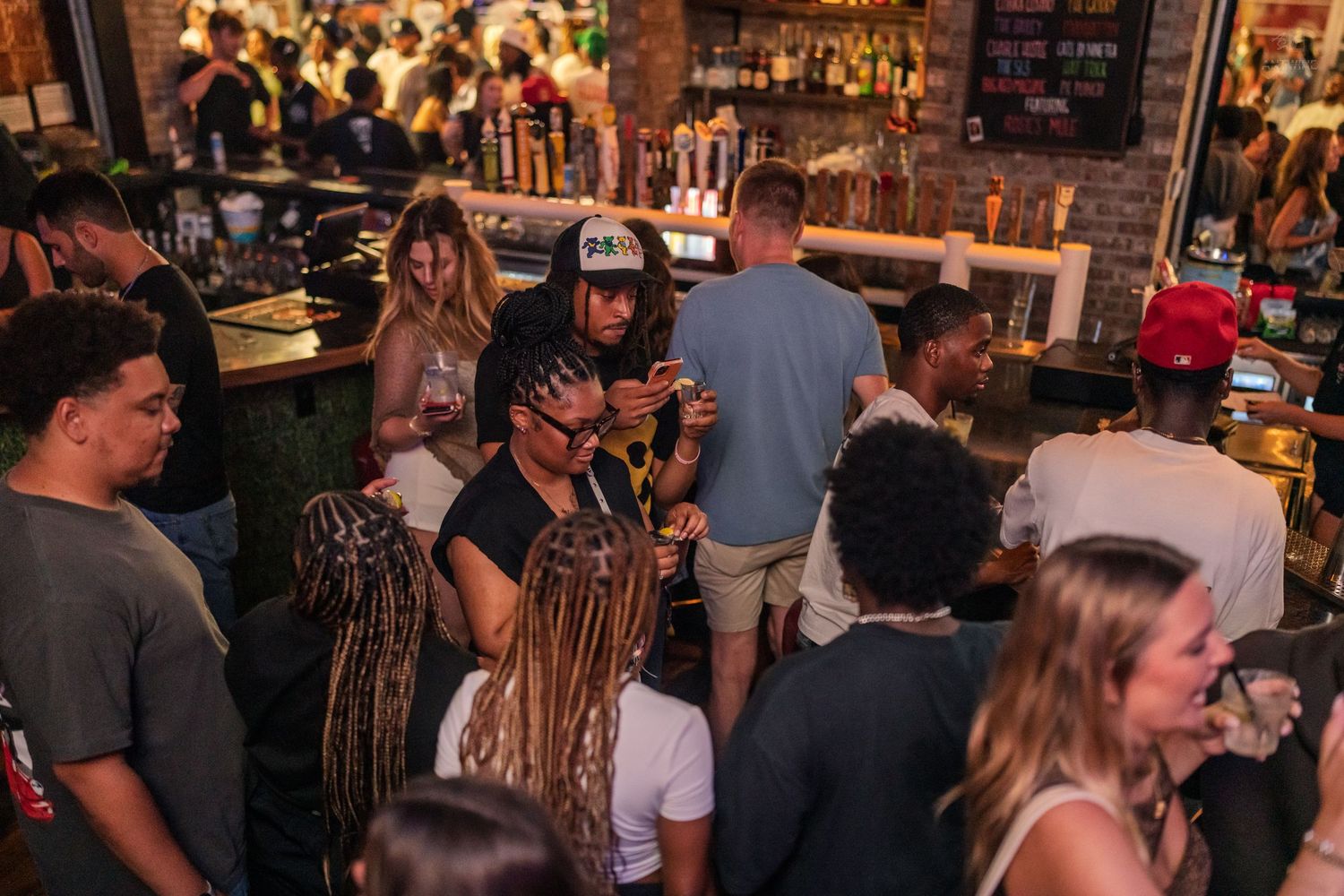 Busy nightclub patrons socializing and using phones near a well-stocked bar counter.