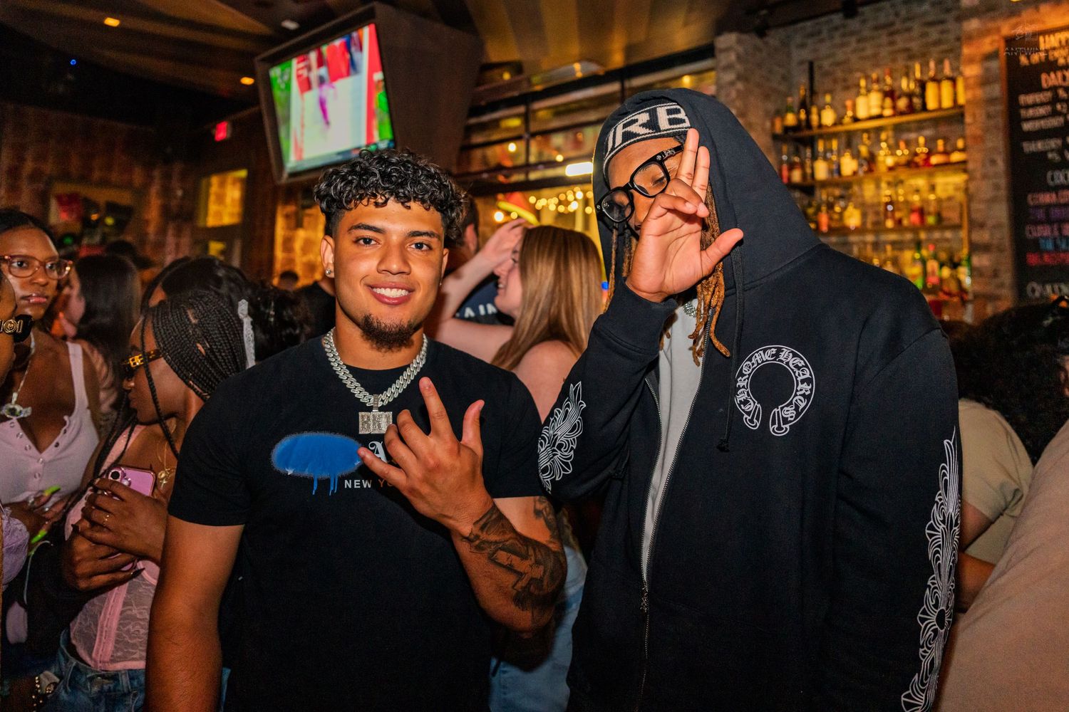 Two men posing and smiling together at a crowded nightclub bar area.