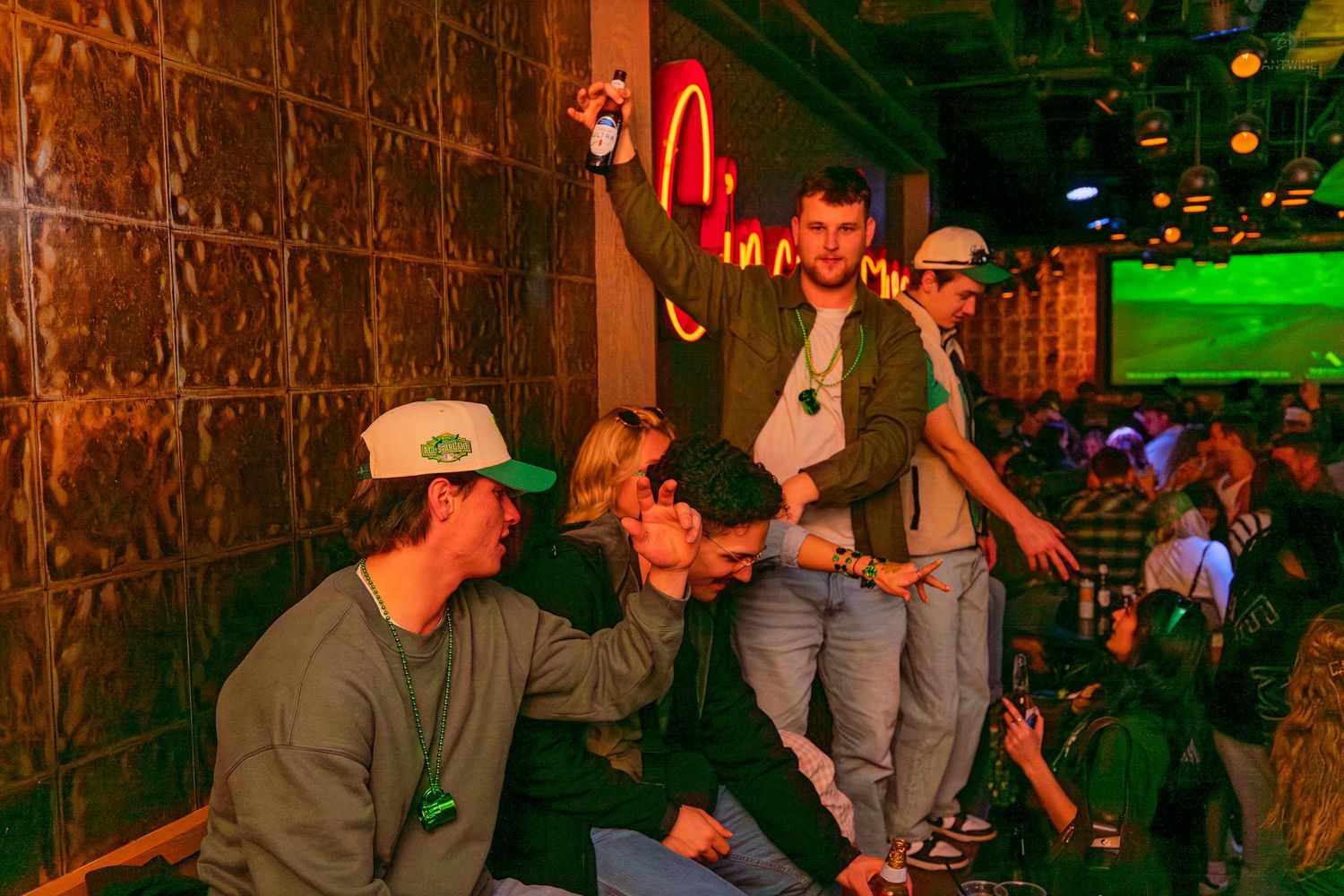 Group of friends posing and celebrating near a neon sign at a green-themed bar party event.