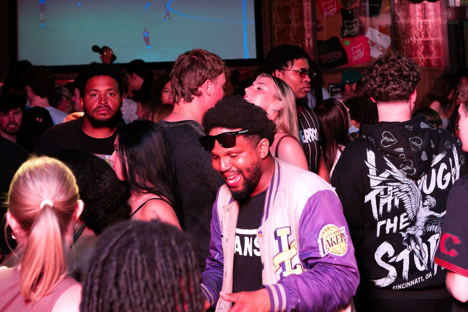 Smiling man in Lakers jacket enjoying a lively crowded nightclub party event.