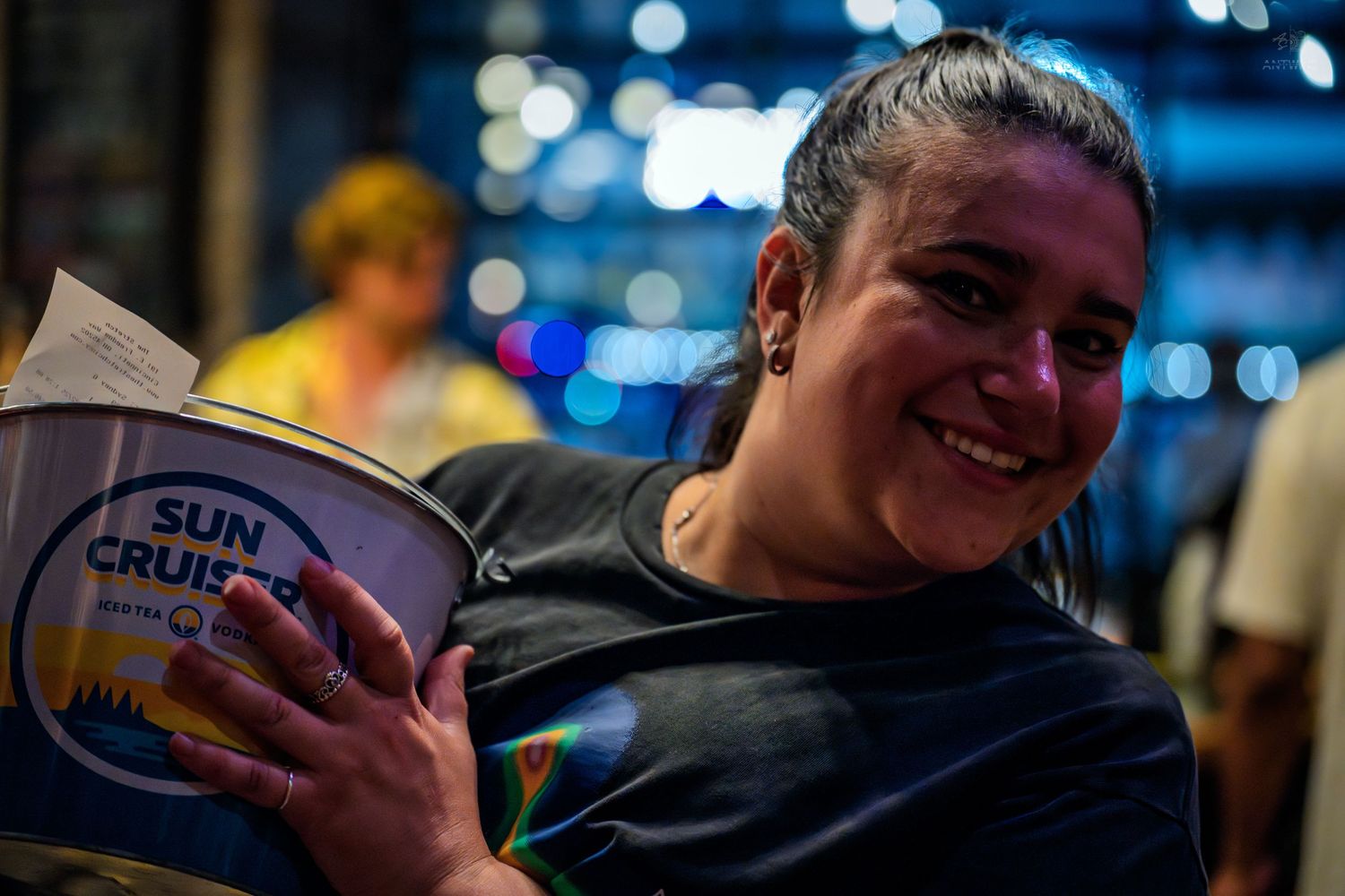 Happy woman clutching a Sun Cruiser branded bucket while smiling at a night event.