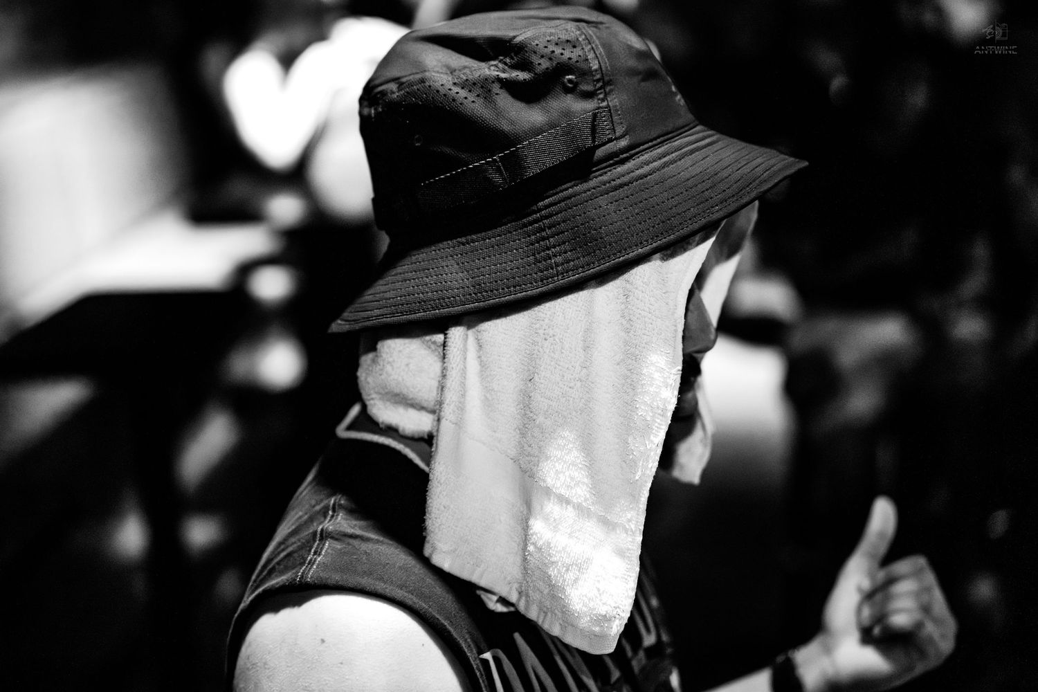 Black and white photo of person wearing bucket hat with towel over face outdoors.