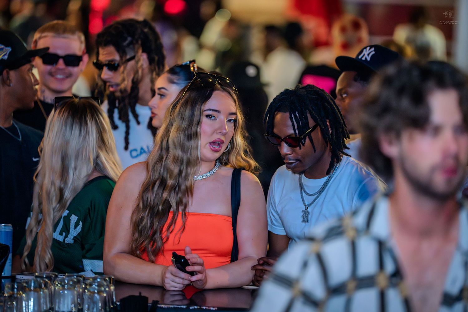 Woman in orange top holding phone surrounded by crowd at busy nightclub.