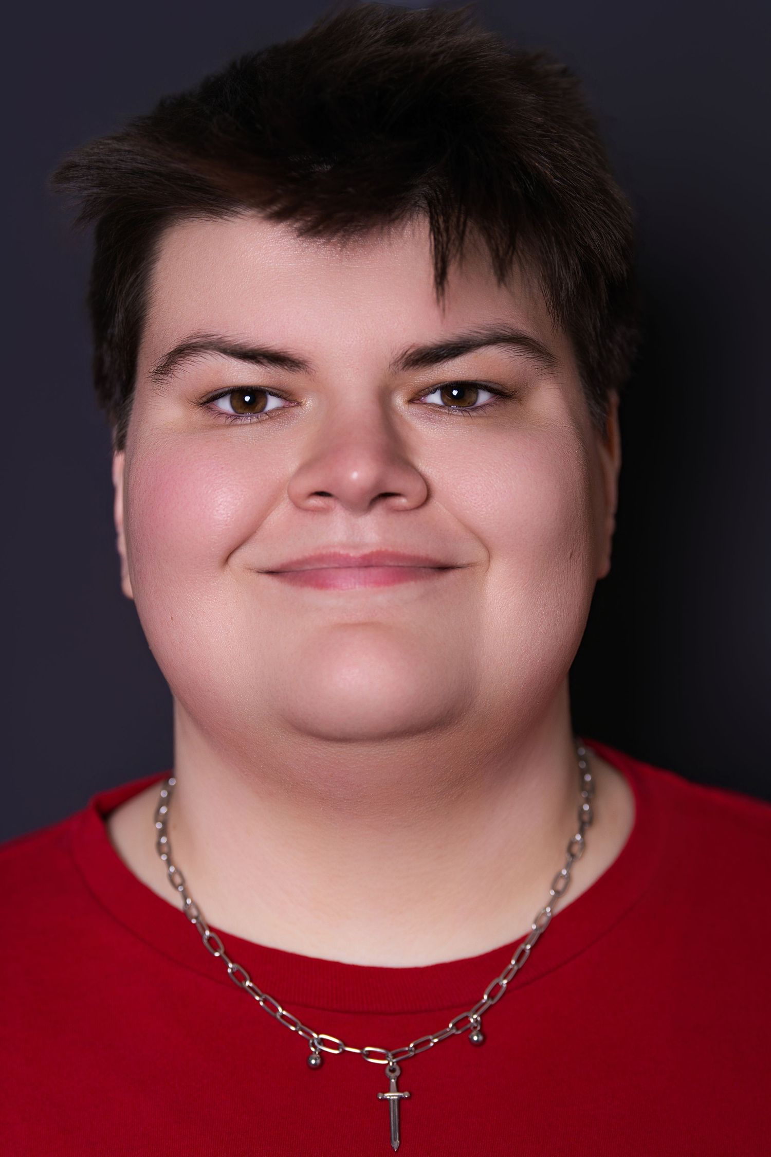 Portrait of a young person in red shirt wearing a silver cross necklace smiling at camera.