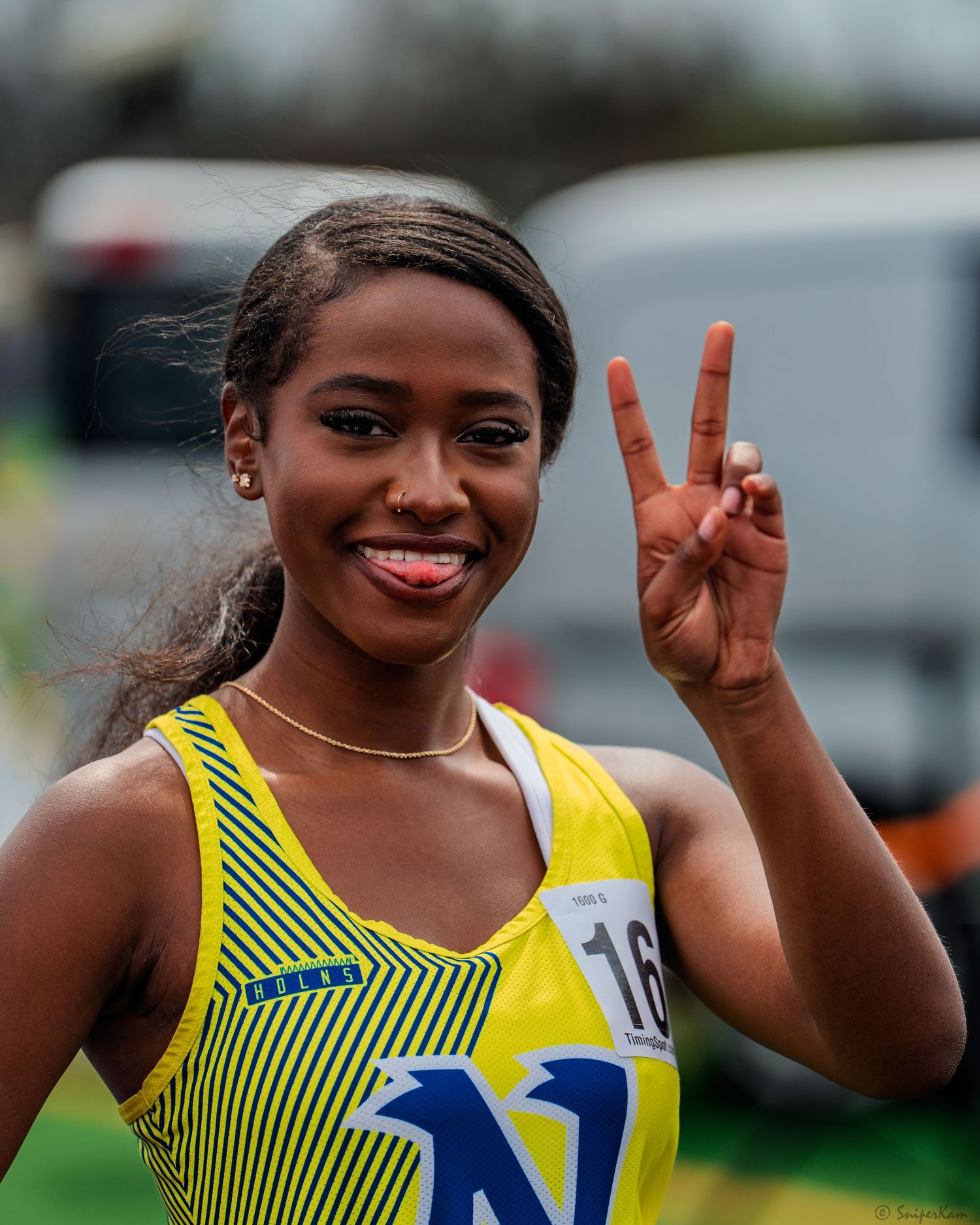 Smiling female track athlete in yellow jersey number 16 gives peace sign at outdoor track meet.