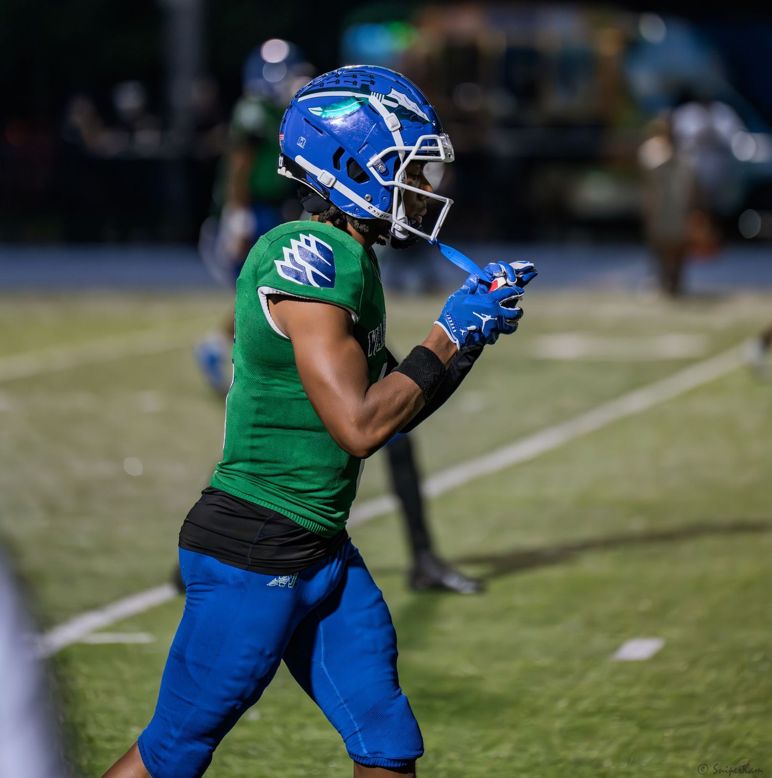 High school football player in green and blue uniform celebrates on field during night game.