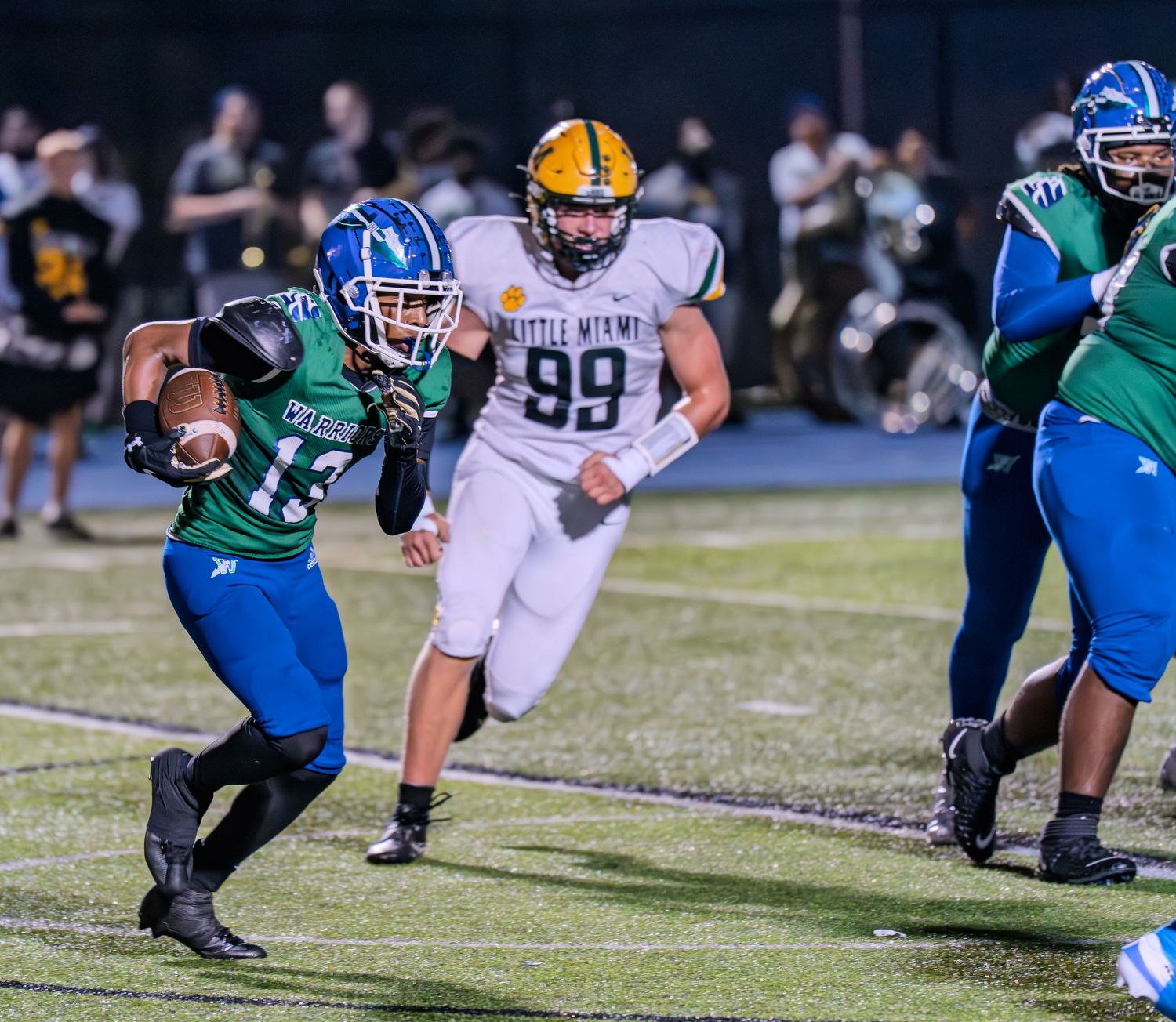 Football player in green uniform runs with ball while being chased by defender number 99 in white.